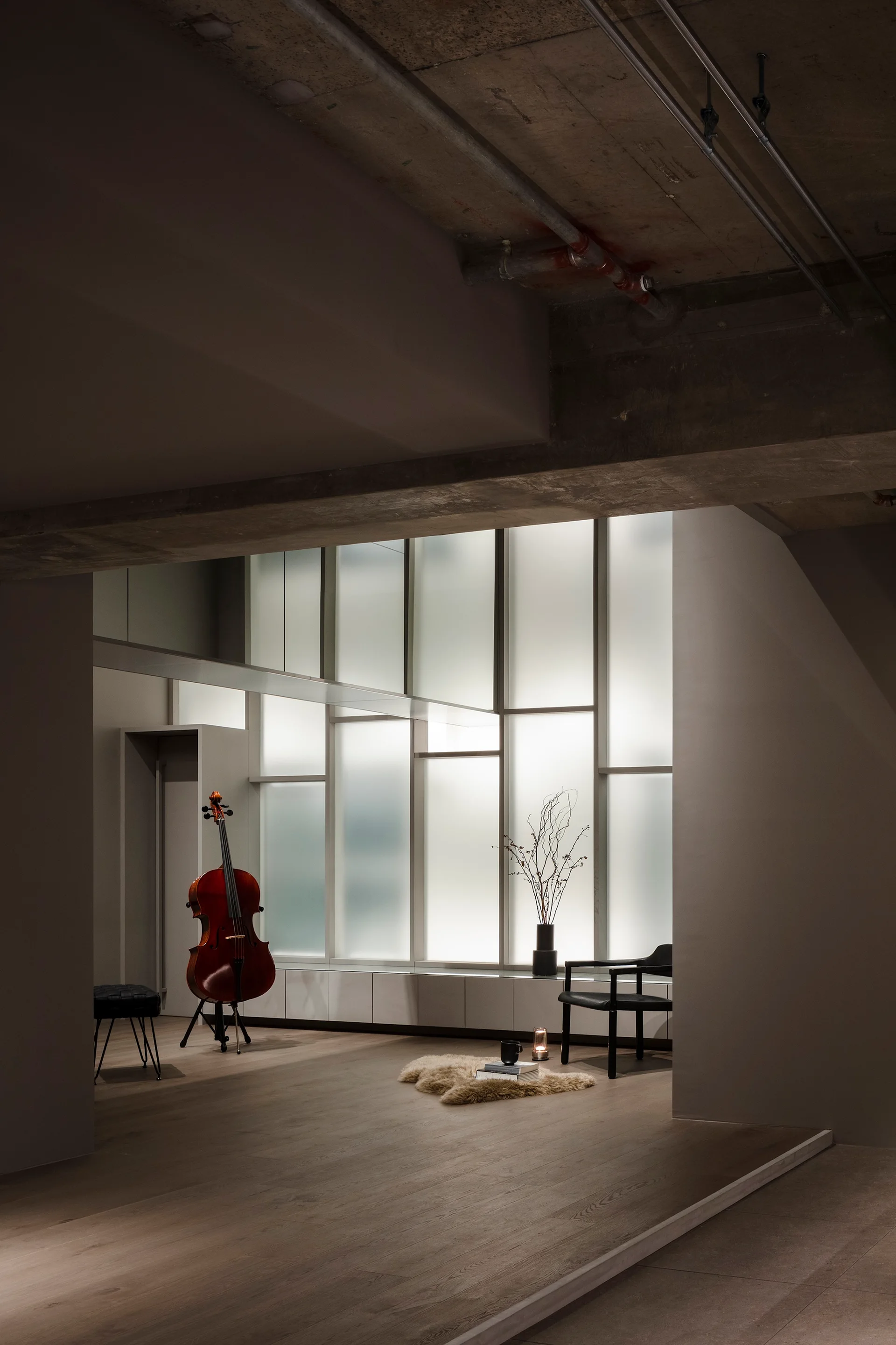 A minimalist room with a cello, black armchair, and sheepskin rug set against a large frosted glass window wall under a raw concrete ceiling.