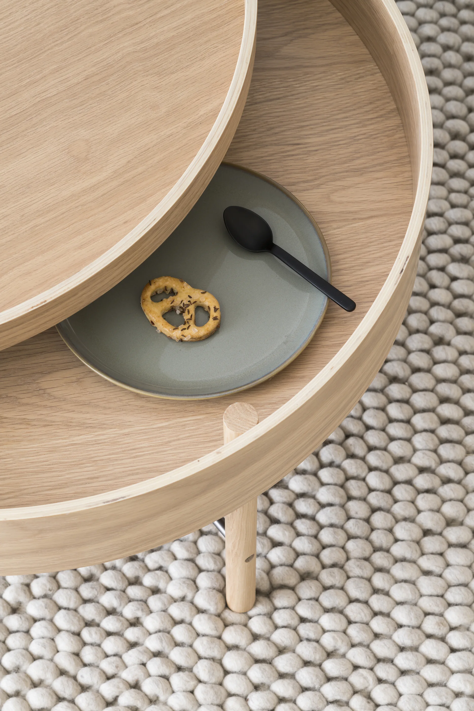 Top-down view of a tiered light wood coffee table with a grey plate on the lower level, resting on a textured white bobble rug.
