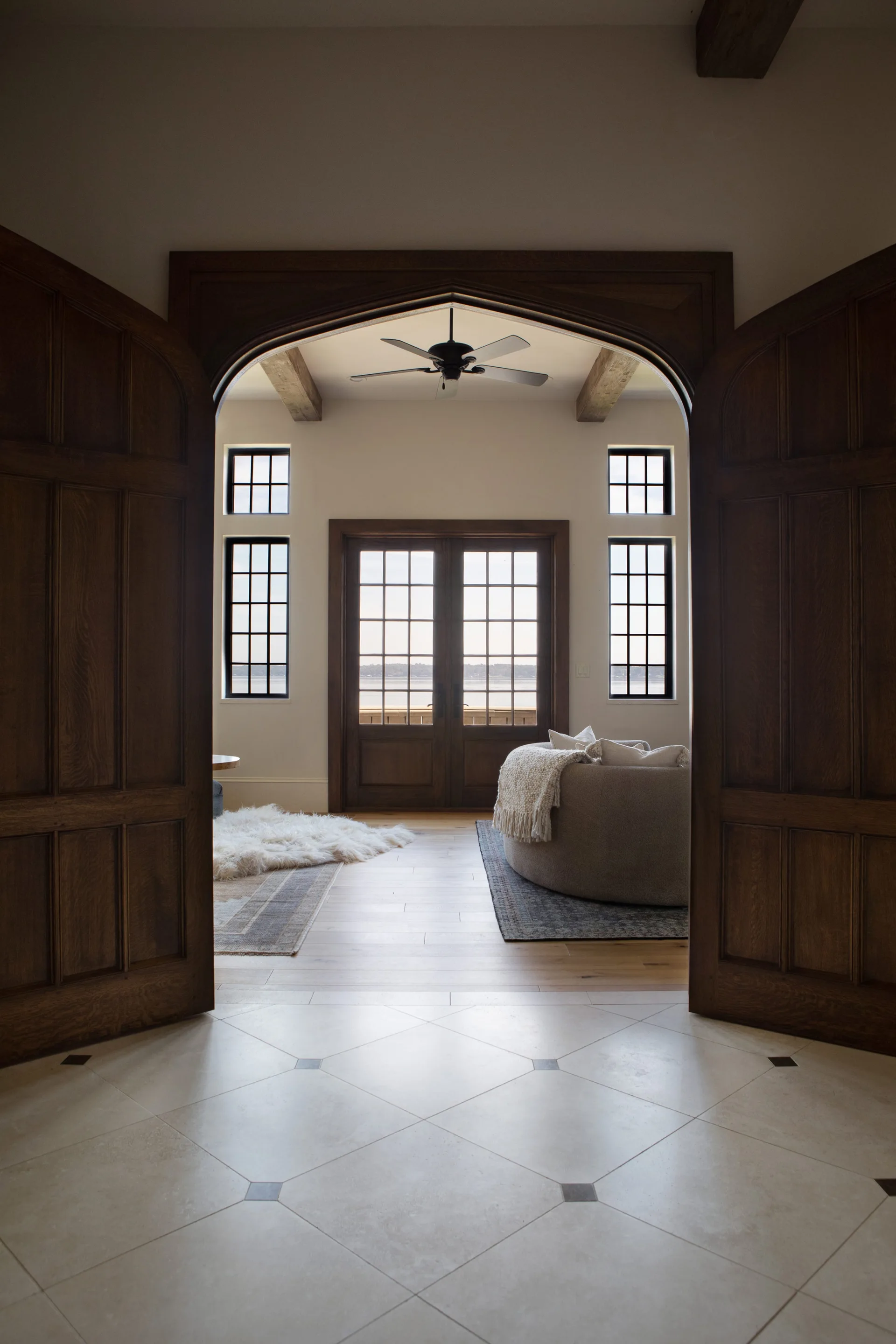 A view through a dark wood arched entryway into a bright living room with a curved sofa, exposed ceiling beams, and French doors.