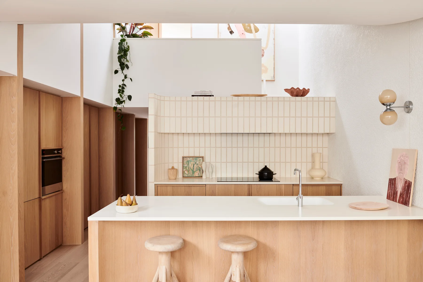 A light wood kitchen features a central island with two matching wooden stools, a white countertop with a sink, and a vertically tiled cream backsplash.