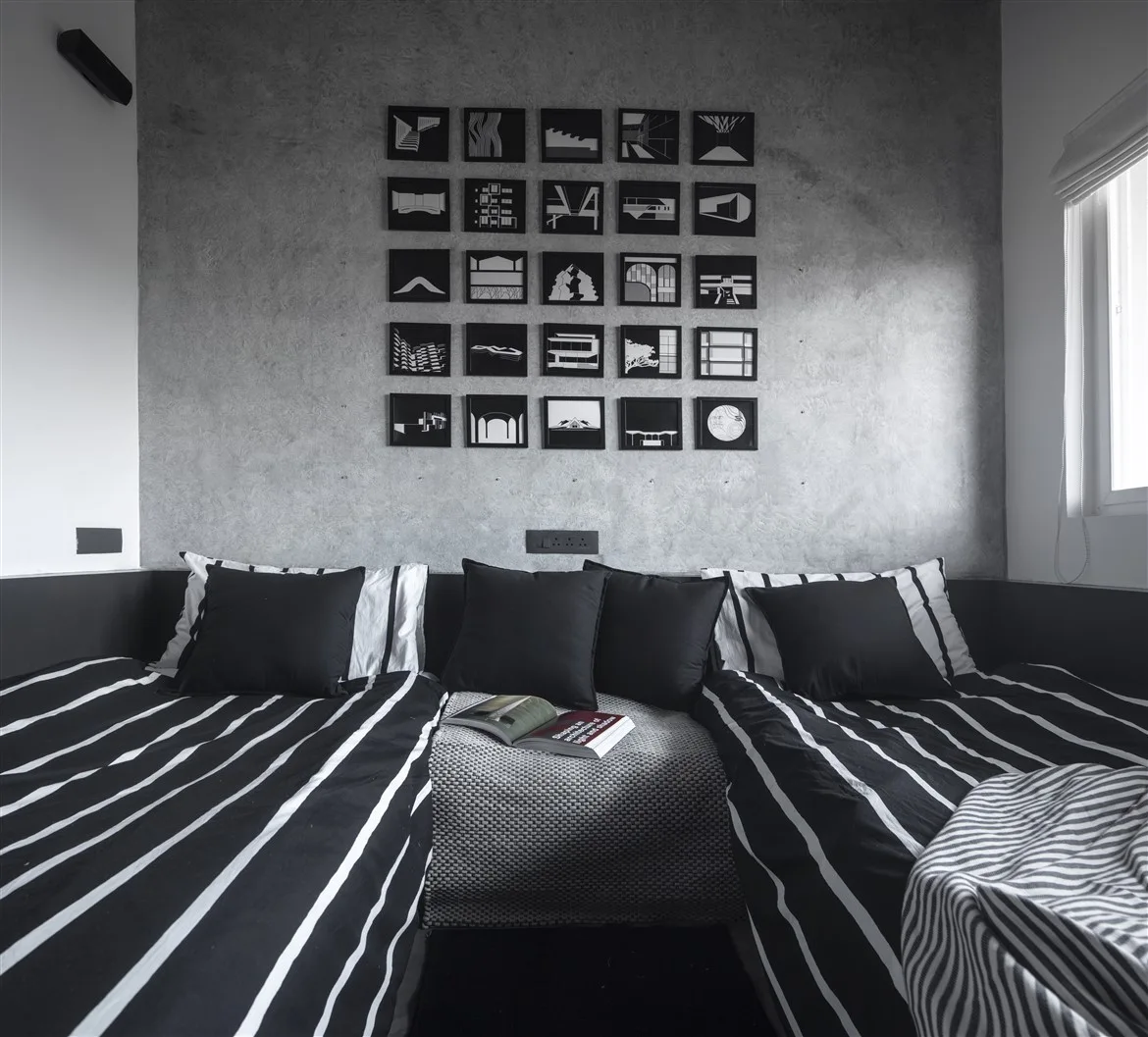Symmetrical twin beds with black and white striped bedding sit against a textured grey concrete wall featuring a square grid gallery of black-framed prints.