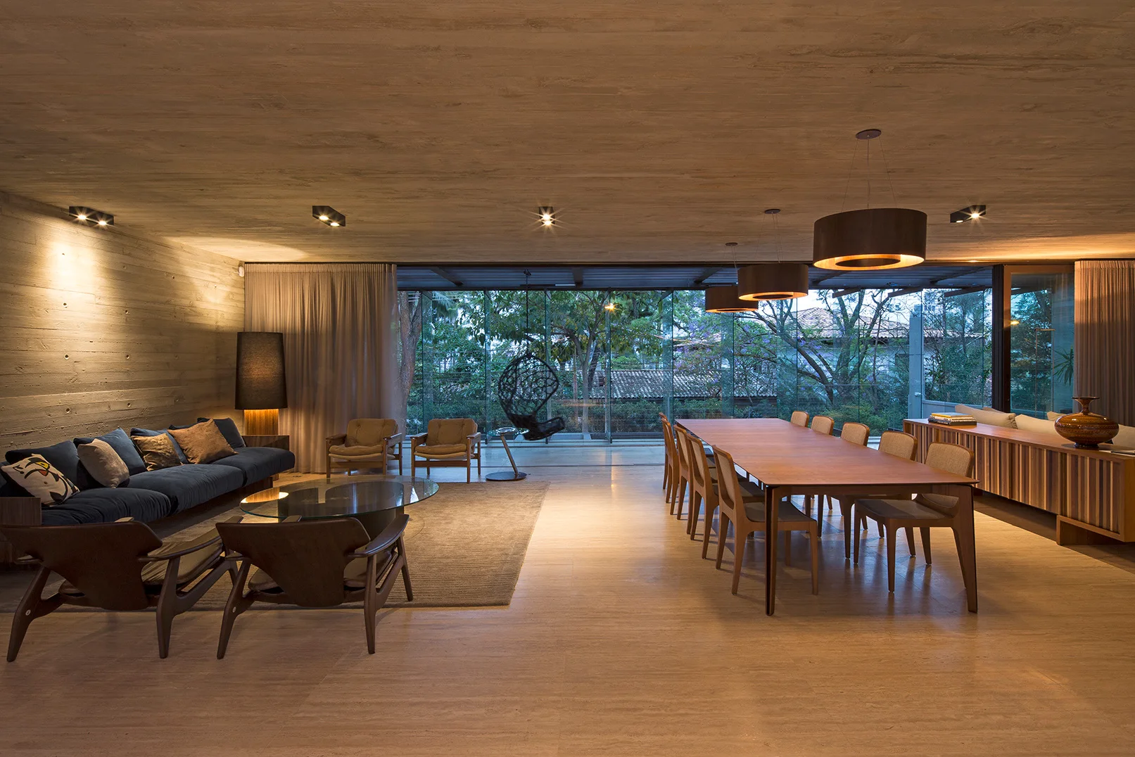 An open-plan living and dining room with a blue sofa, a long wood dining table, exposed concrete walls, and large glass doors looking onto a garden.