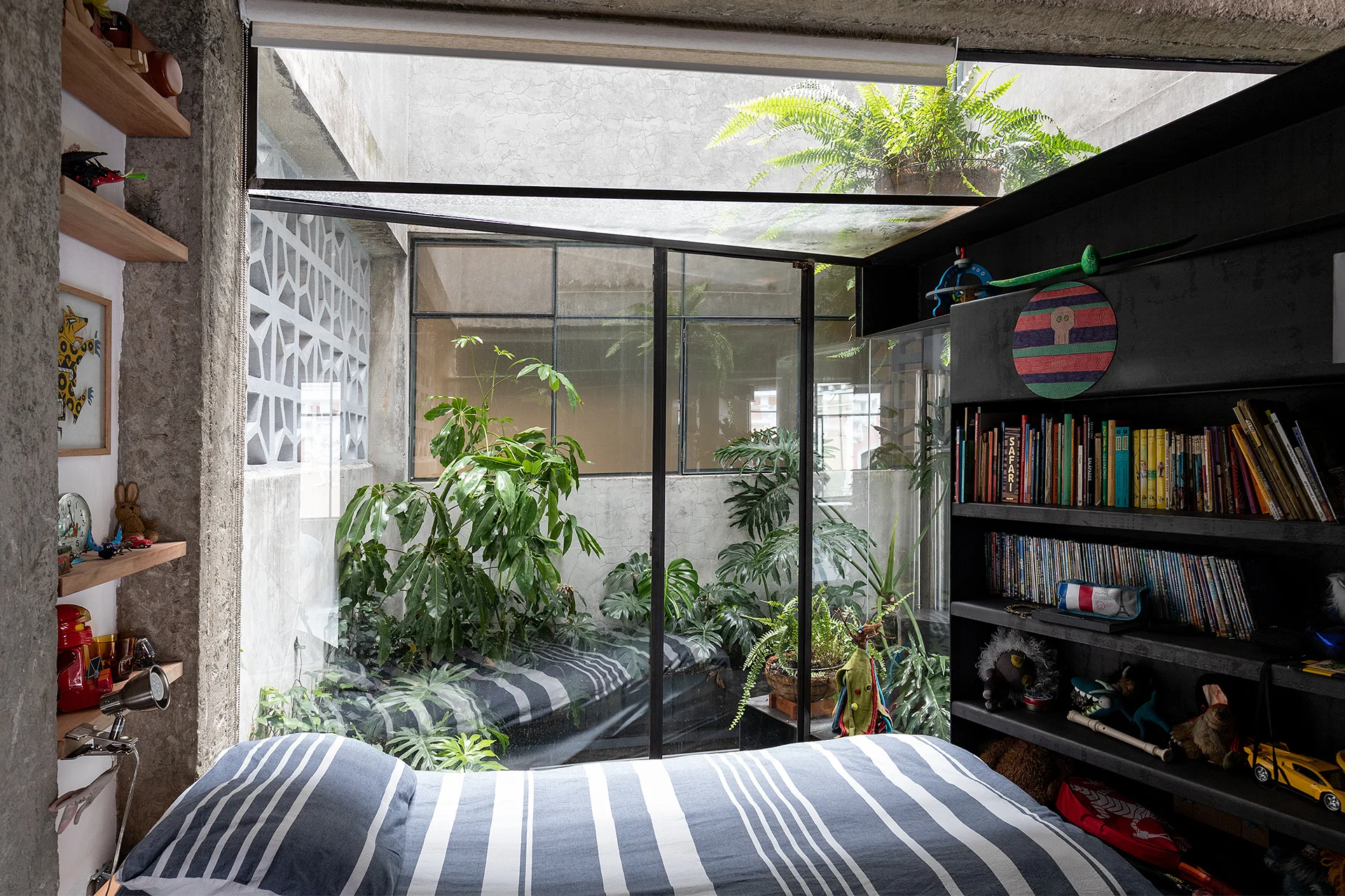 An industrial bedroom features a bed with striped blue sheets next to a large glass partition showing a plant-filled light well and raw concrete walls.