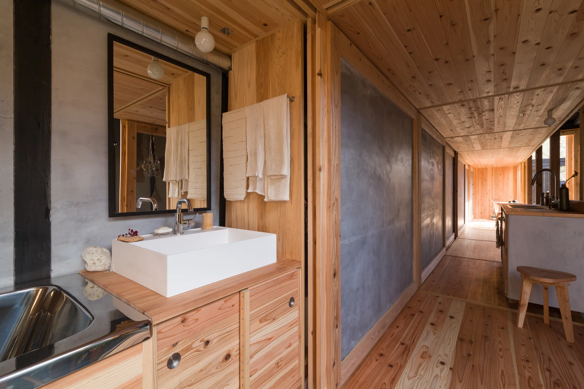 A bathroom vanity with a white vessel sink and black-framed mirror set against concrete walls and pine wood panels next to a hallway.