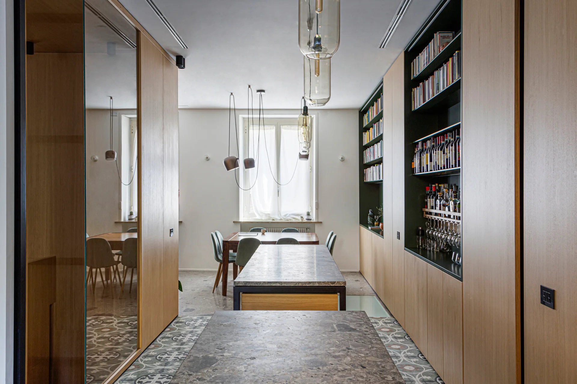 A dining room features built-in wood cabinets, a stone island, glass pendant lights, and a transition from patterned tiles to a smooth stone floor.