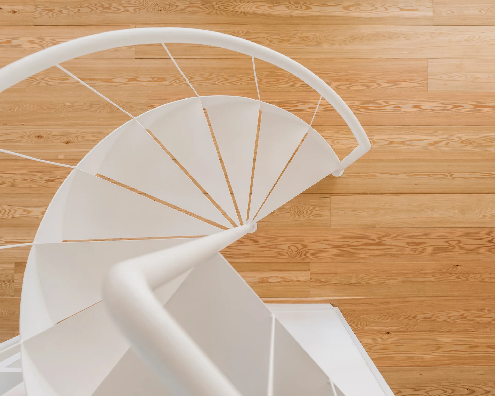 A high-angle view of a minimalist white metal spiral staircase positioned over warm light brown wood plank flooring.