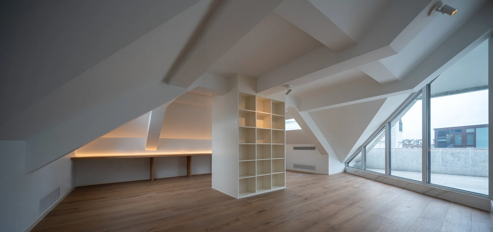 A minimalist attic room features a central white bookshelf, a long built-in wood desk under a sloped ceiling, and a large angular glass window.