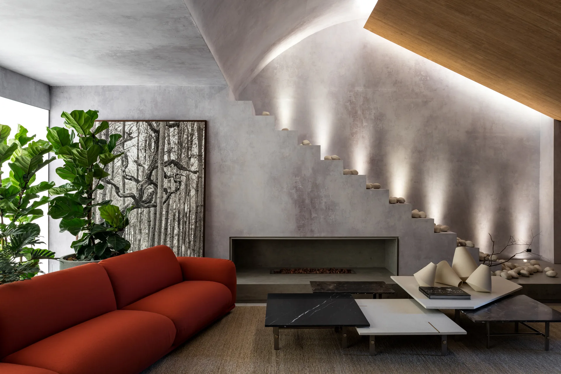 A contemporary living room features a bright red sofa, nested stone coffee tables, and a sculptural concrete staircase lit by recessed wall uplights.