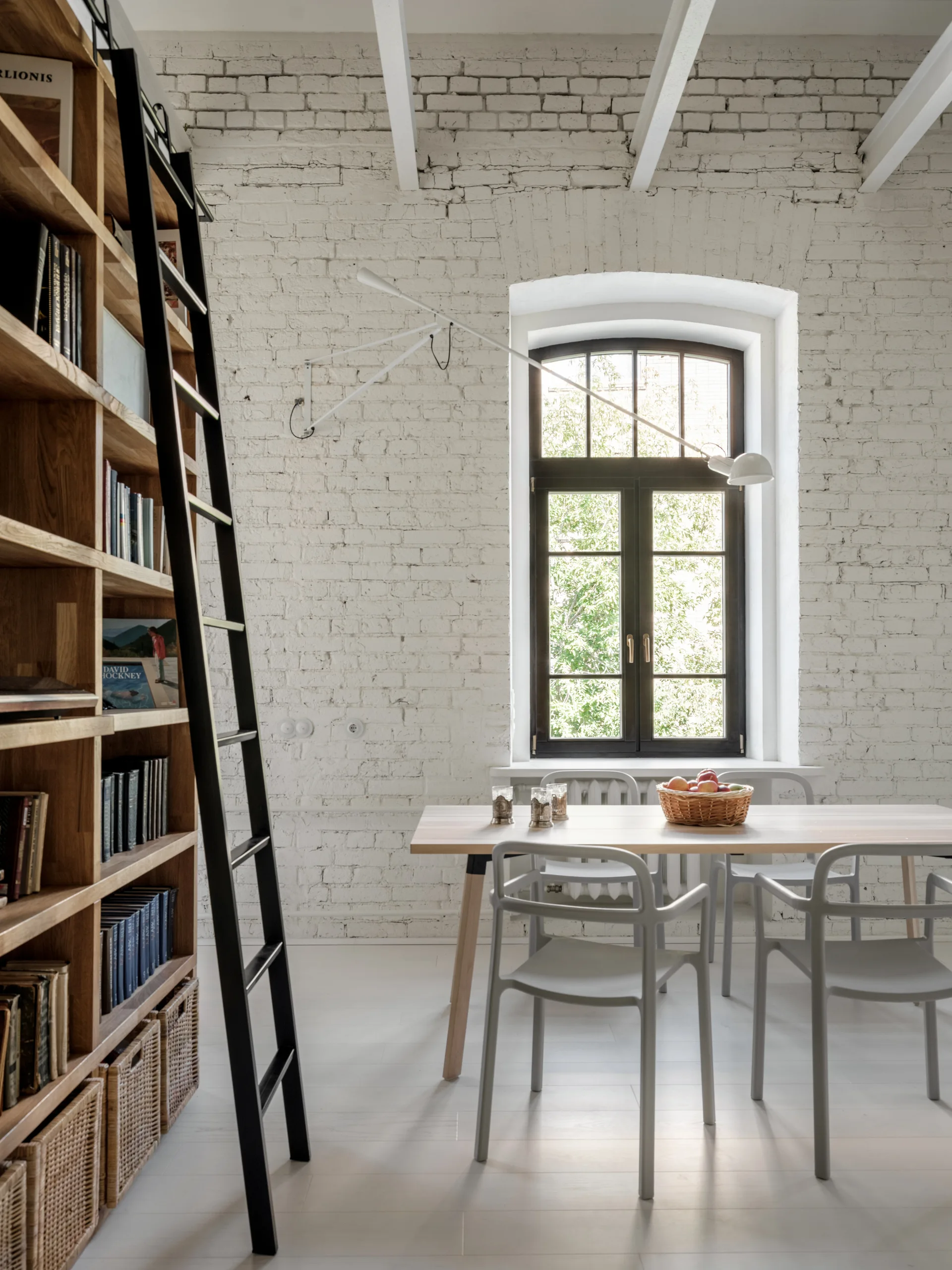 A light wood dining table with grey chairs sits next to a tall wooden bookcase with a black rolling ladder against a white painted brick wall.