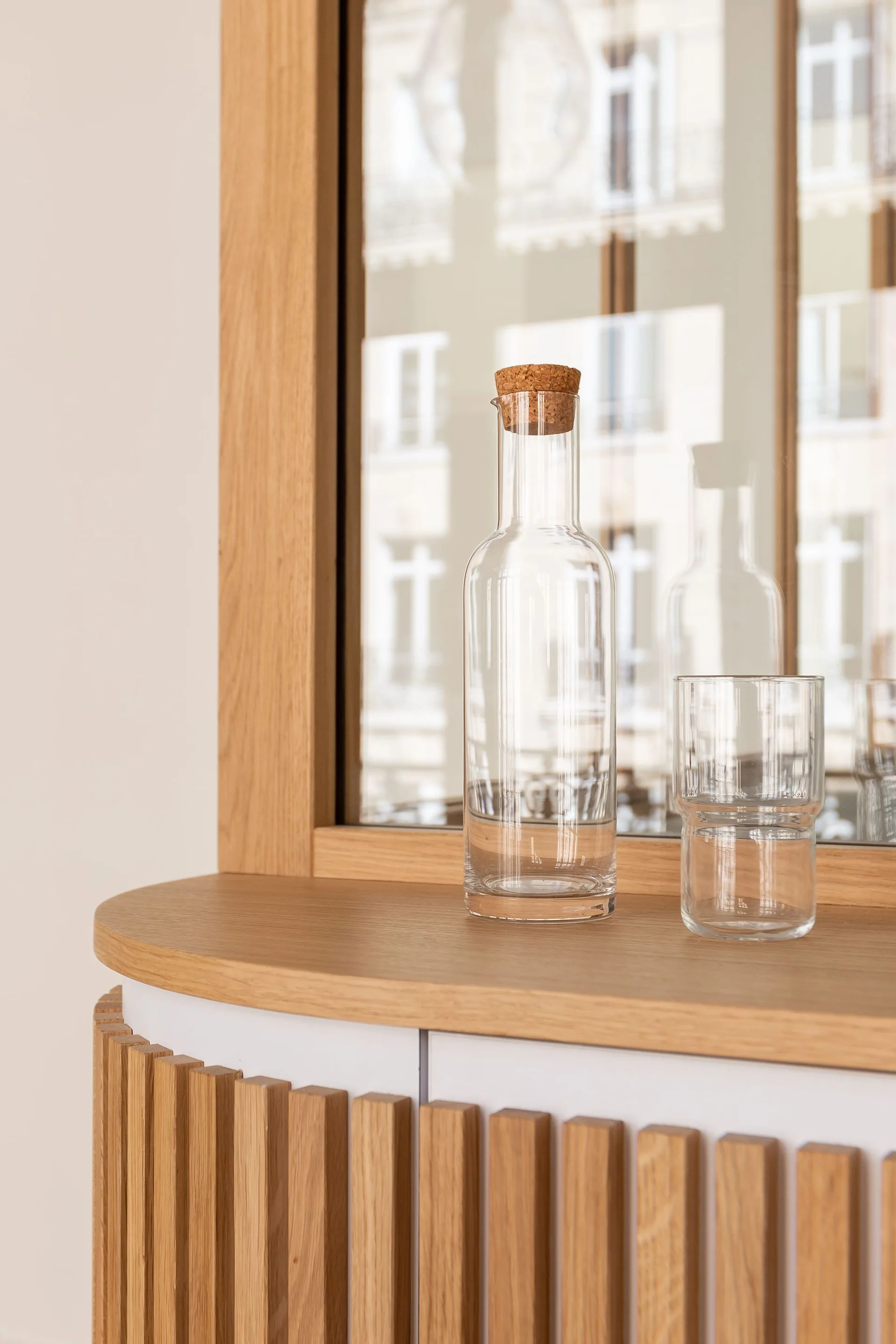 A clear glass decanter and tumbler sit on a light oak curved sideboard with vertical wood slats and a mirrored back.