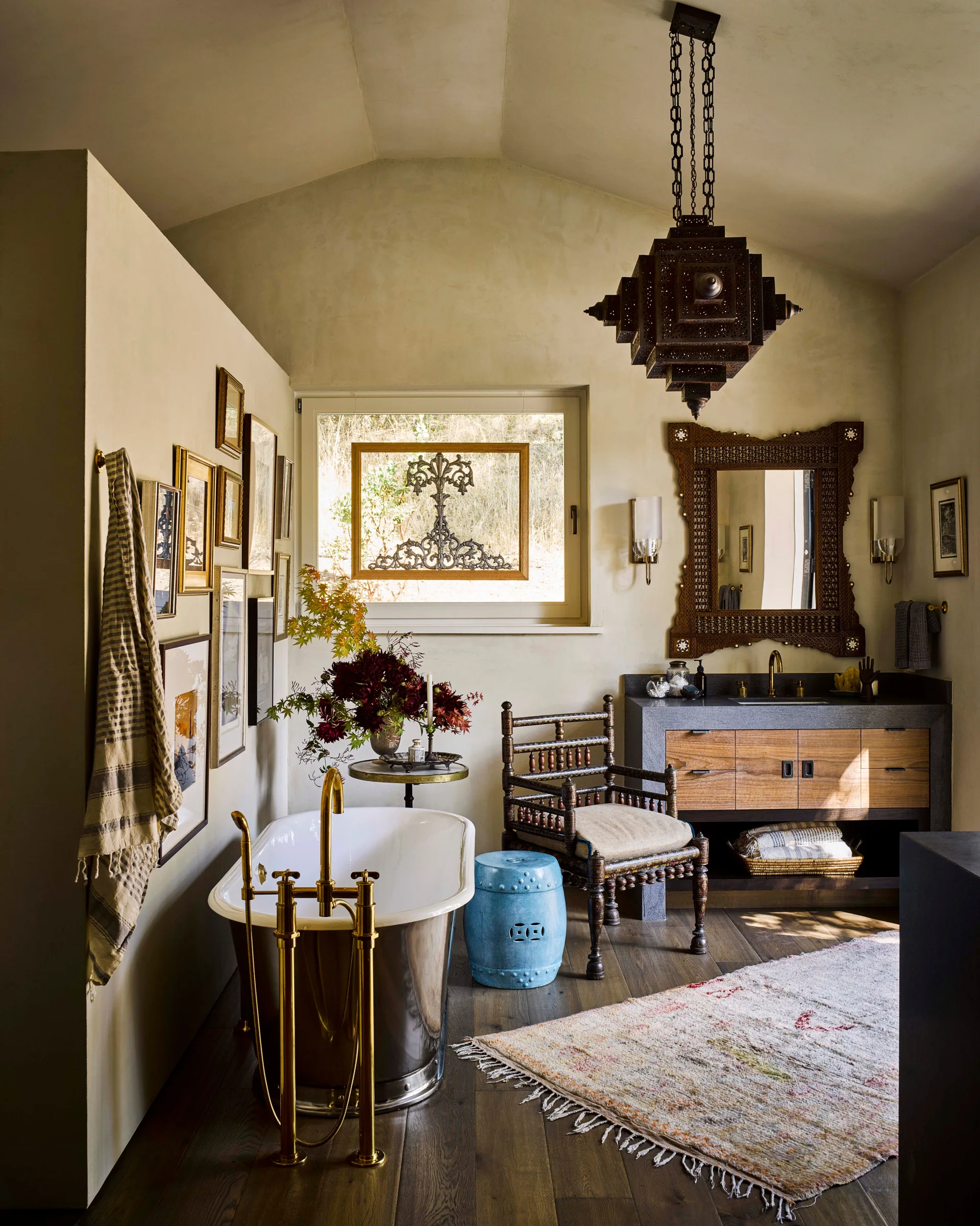 An eclectic bathroom with a metallic freestanding tub, a gallery wall, a rustic spindle chair, and a wood vanity under a vaulted ceiling.