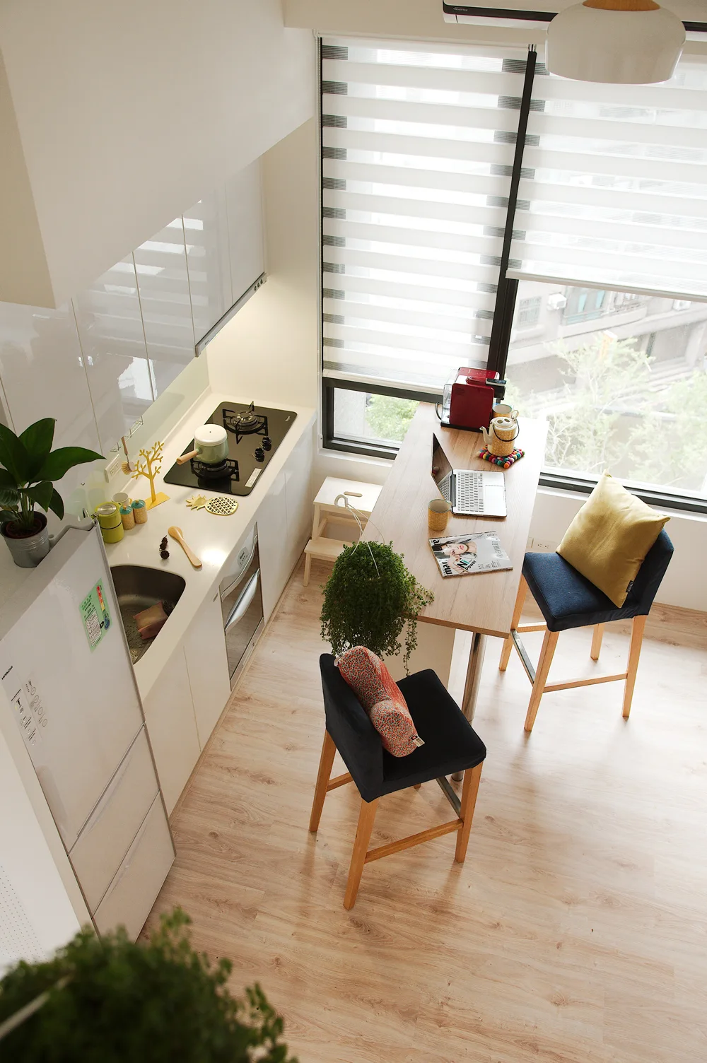 A high-angle view of a compact white kitchen with glossy cabinets, a light wood bar table, navy blue upholstered stools, and striped window shades.
