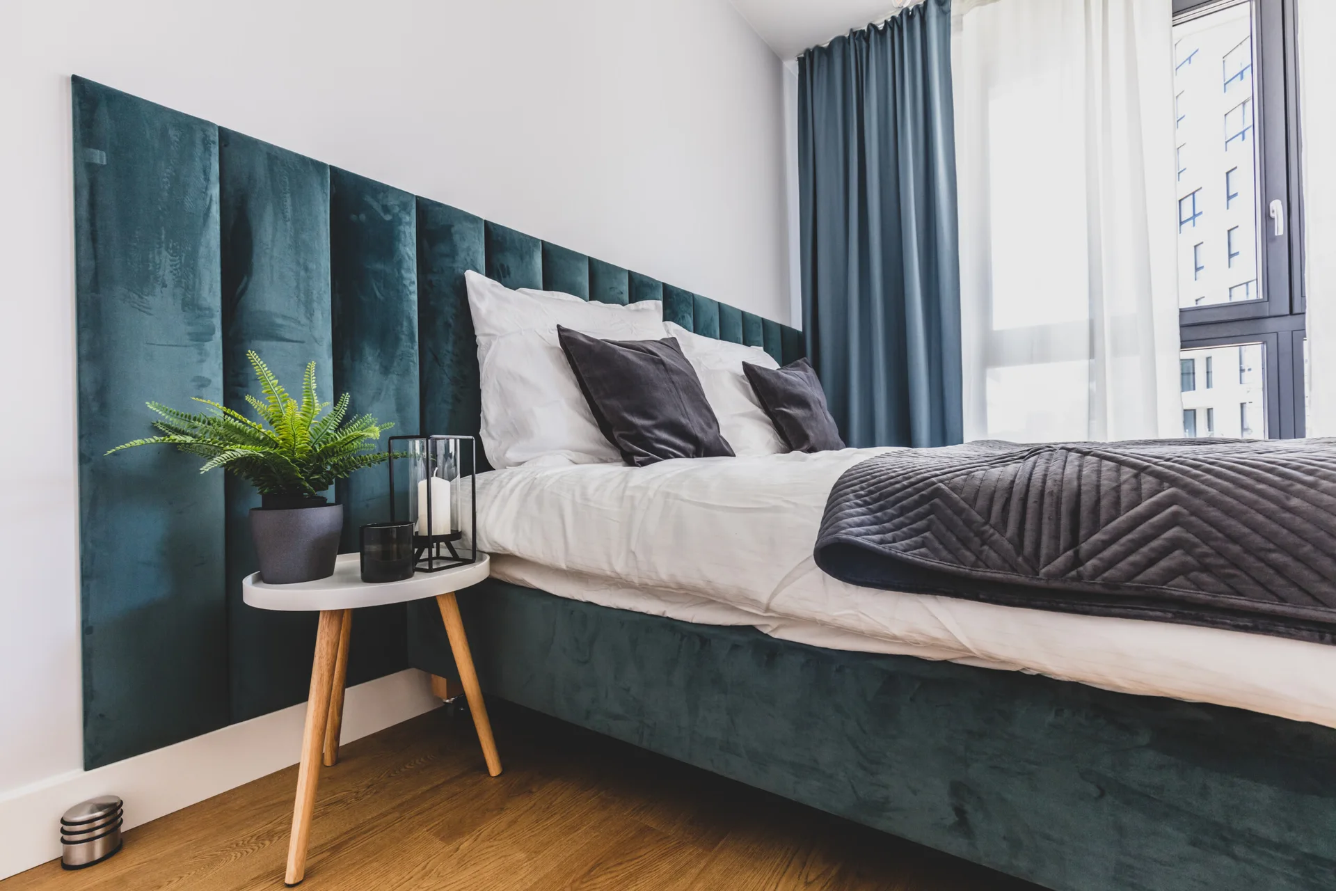 A modern bedroom features a teal velvet upholstered headboard, a white bedside table with a small plant, and matching floor-length curtains.