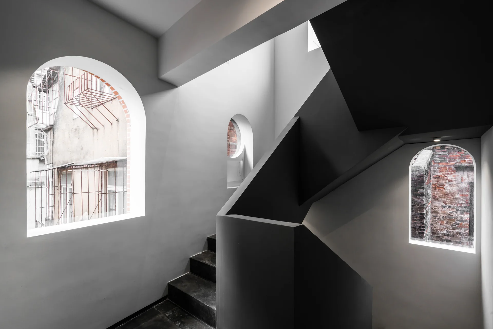 A geometric charcoal staircase rises next to smooth white walls with three distinct arched windows showcasing exterior brick views.