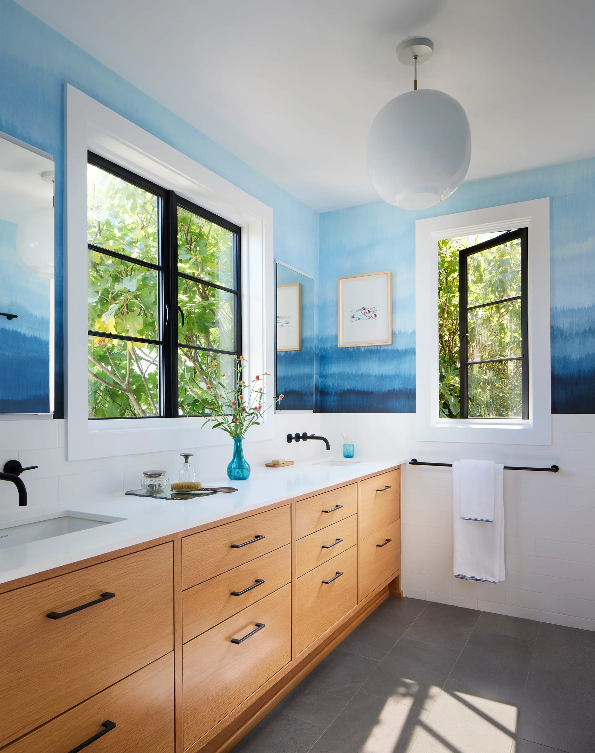 A bright bathroom features a light wood double vanity with white countertops, blue ombre landscape wallpaper, and black-framed windows.