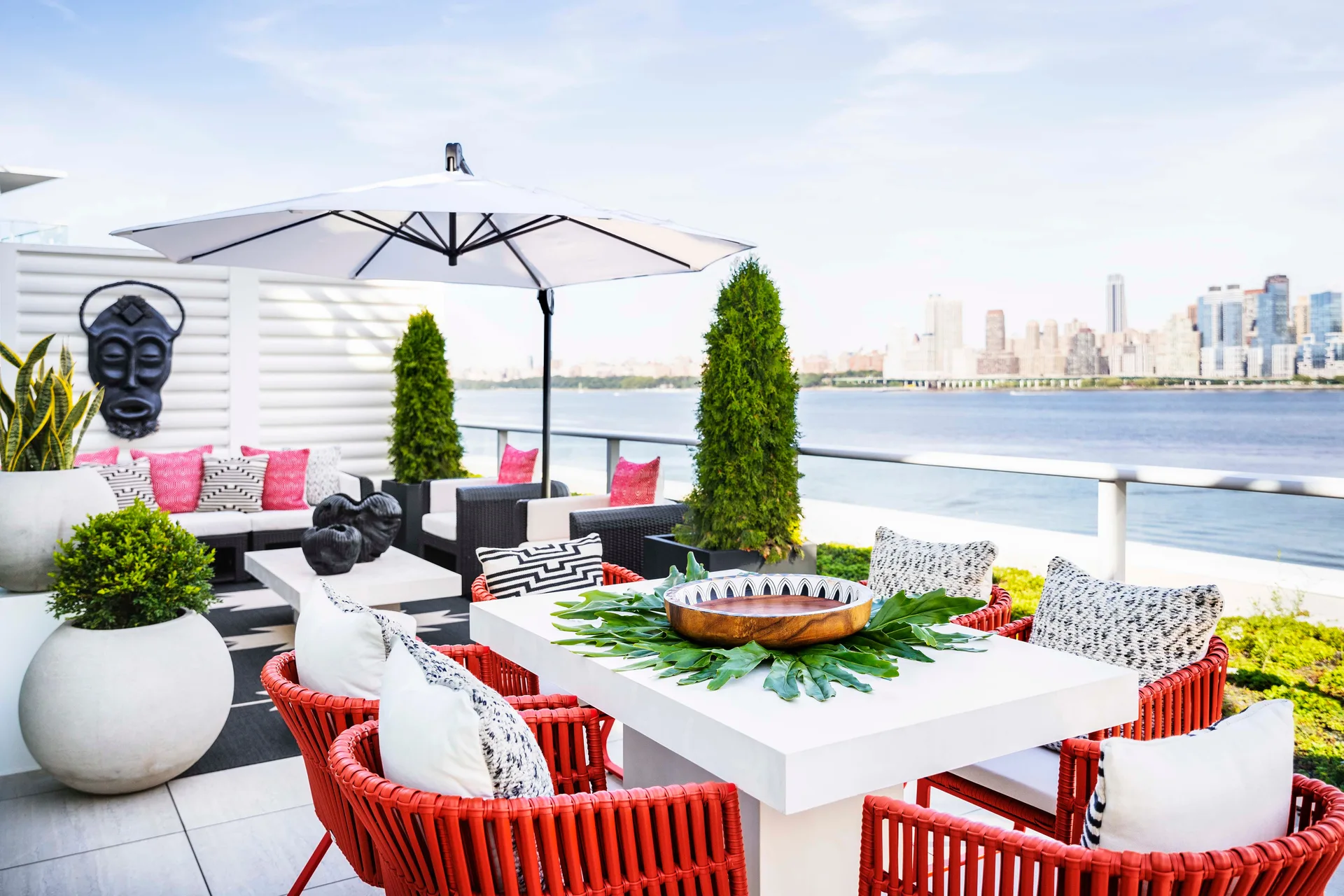 A modern rooftop terrace features red woven dining chairs, a white table, a large umbrella, and a low-profile sofa overlooking a city skyline and water.