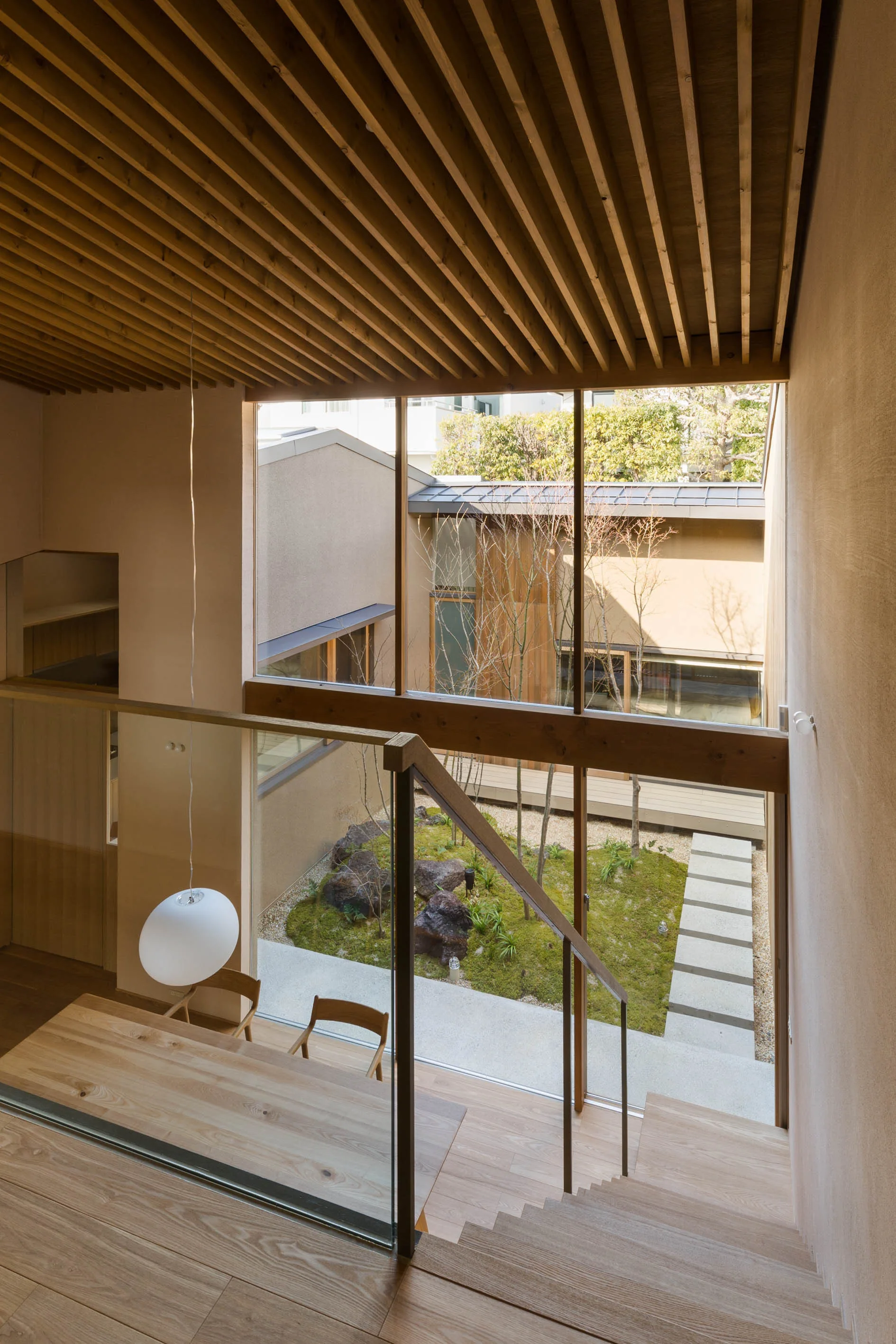 A wooden staircase with a glass railing looks out onto an interior courtyard garden with moss and bare trees through a large window.