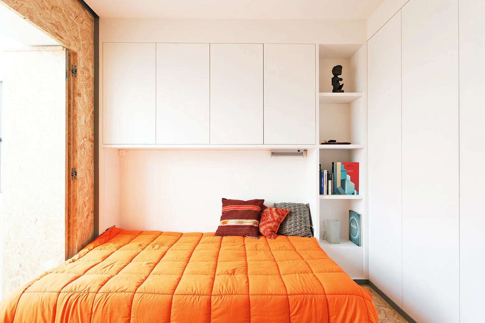 A minimalist bedroom features a bed with orange quilted bedding tucked under white built-in storage cabinets and an open side bookshelf.