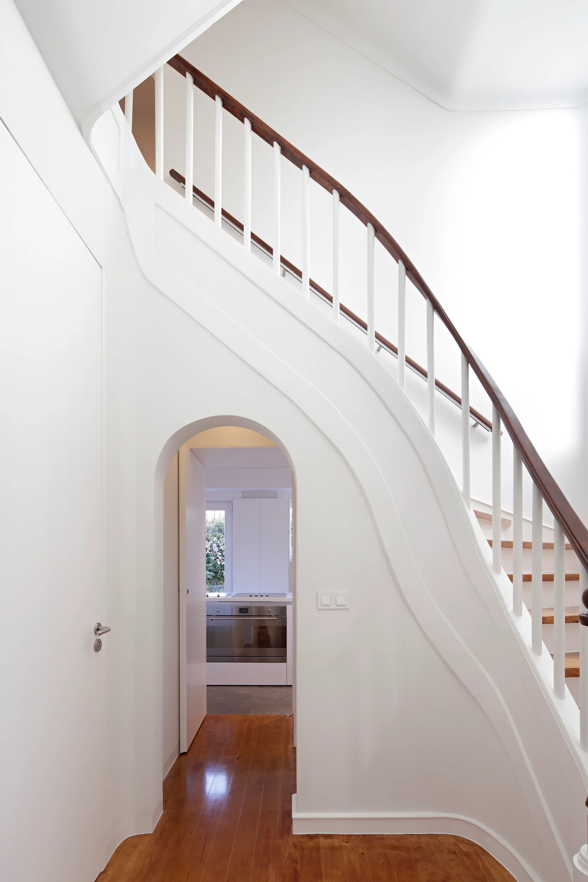 A curved white staircase with white balusters and a dark wood handrail features an arched doorway tucked underneath leading to a kitchen.