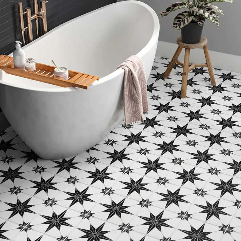 A white freestanding bathtub sits on black and white star-patterned floor tiles next to a wooden stool holding a potted plant.