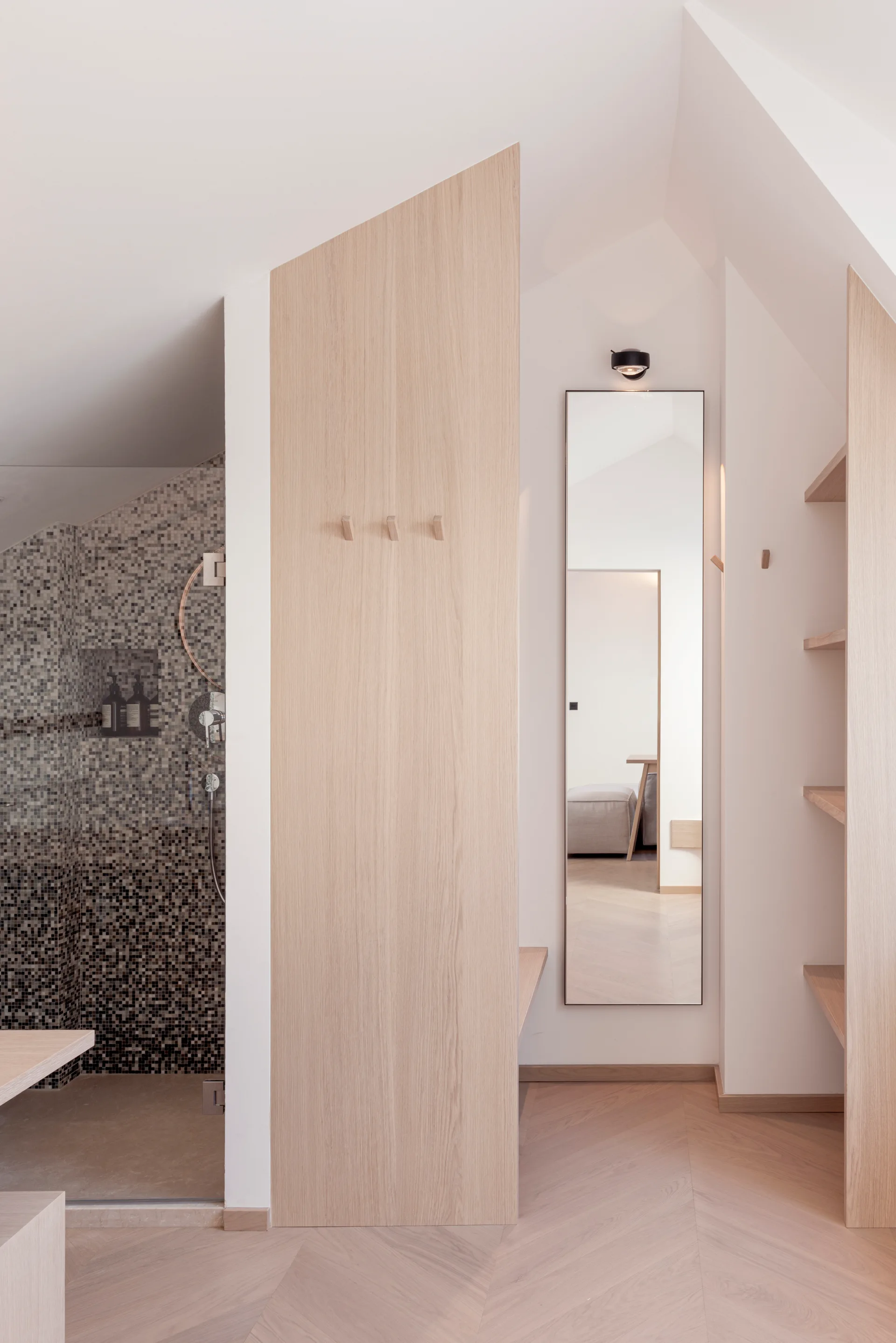 A minimalist dressing area featuring a light wood partition wall, built-in shelving, herringbone floors, and a narrow full-length wall mirror.