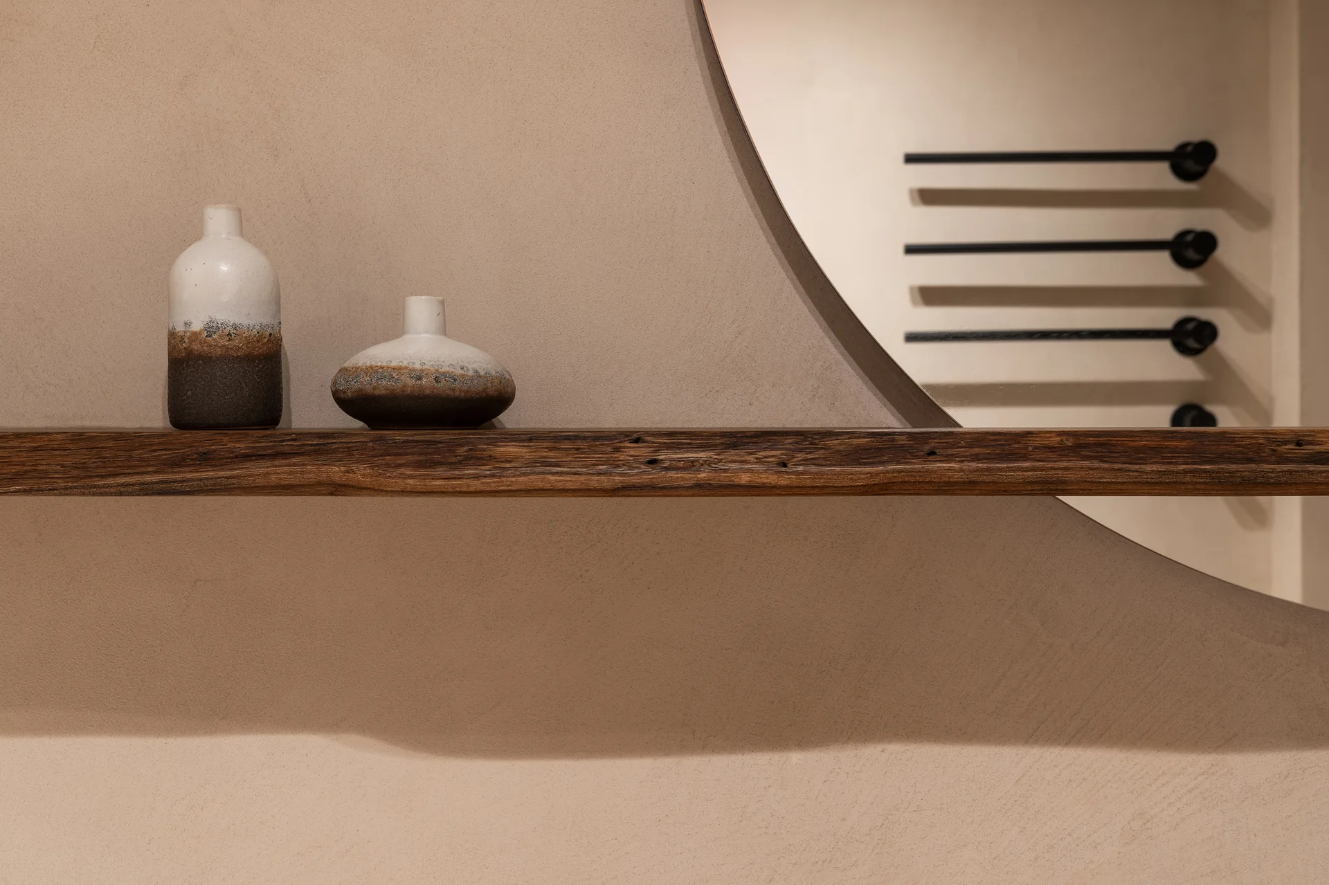 Two ceramic vases sit on a rustic dark wood floating shelf in front of a round wall mirror against a beige plastered wall.