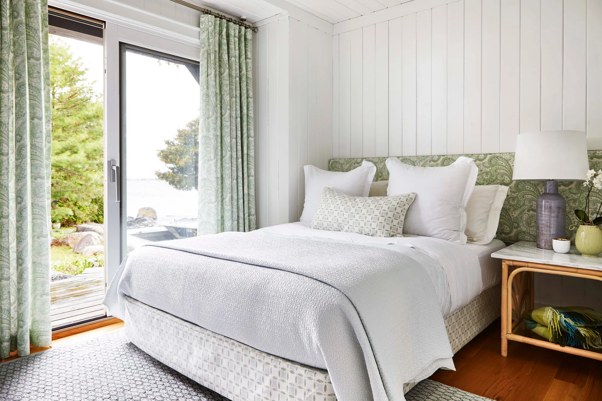 Cottage bedroom with a white bed, patterned sage green curtains next to a sliding glass door, and vertical white wood paneling on the walls.