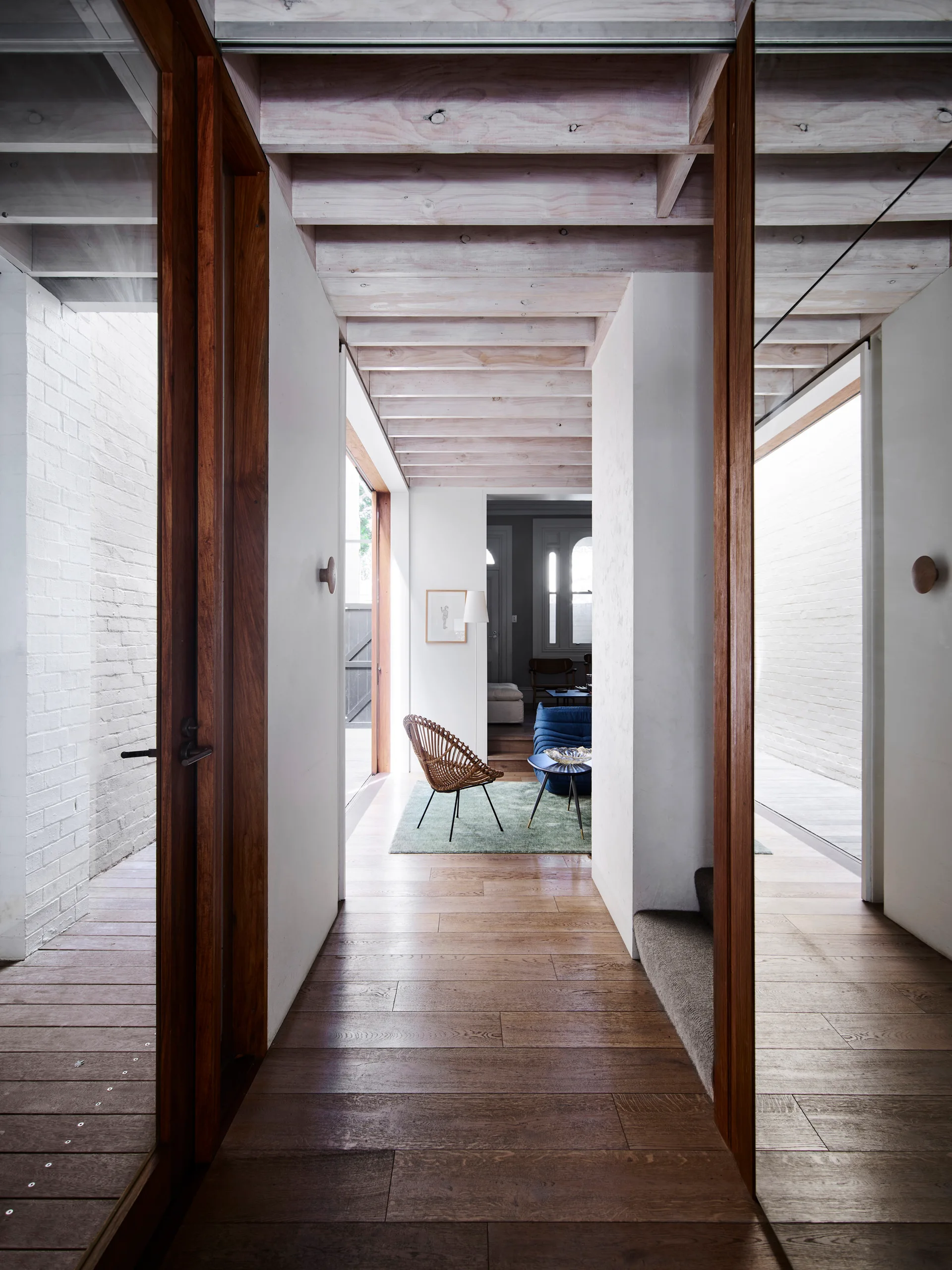 A wooden-floored hallway with exposed ceiling beams leads past a white brick wall toward a sunlit living room with a green rug.