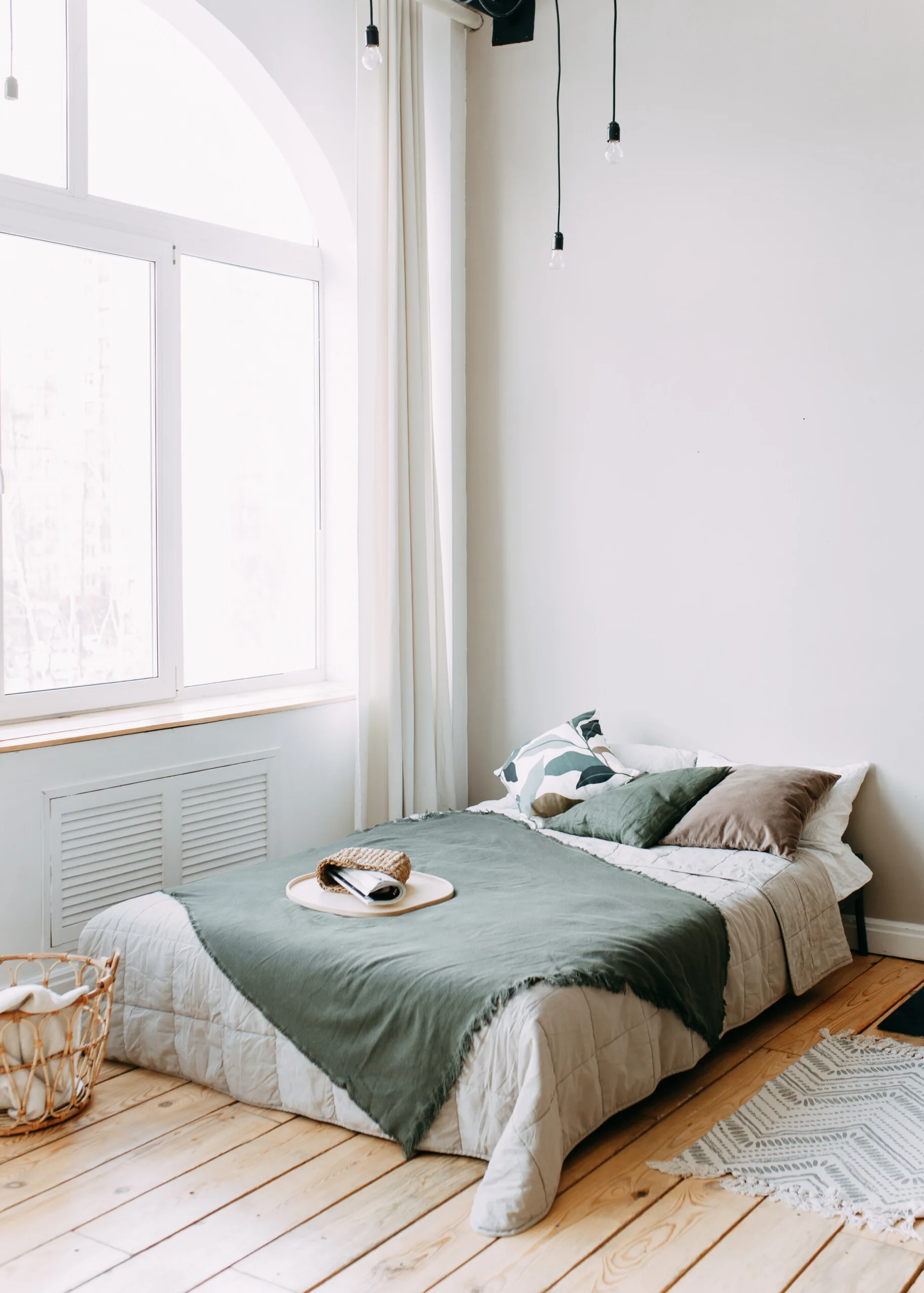 A minimalist bedroom features a low bed with olive green bedding, set beside a large arched window on light wood plank flooring.