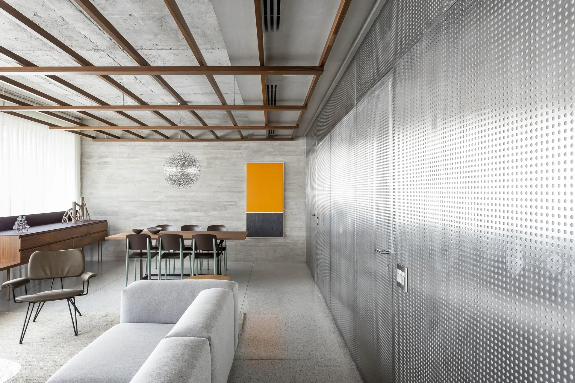 A modern open-plan living and dining area features a perforated silver metal wall, exposed concrete surfaces, and a dark wood grid ceiling.