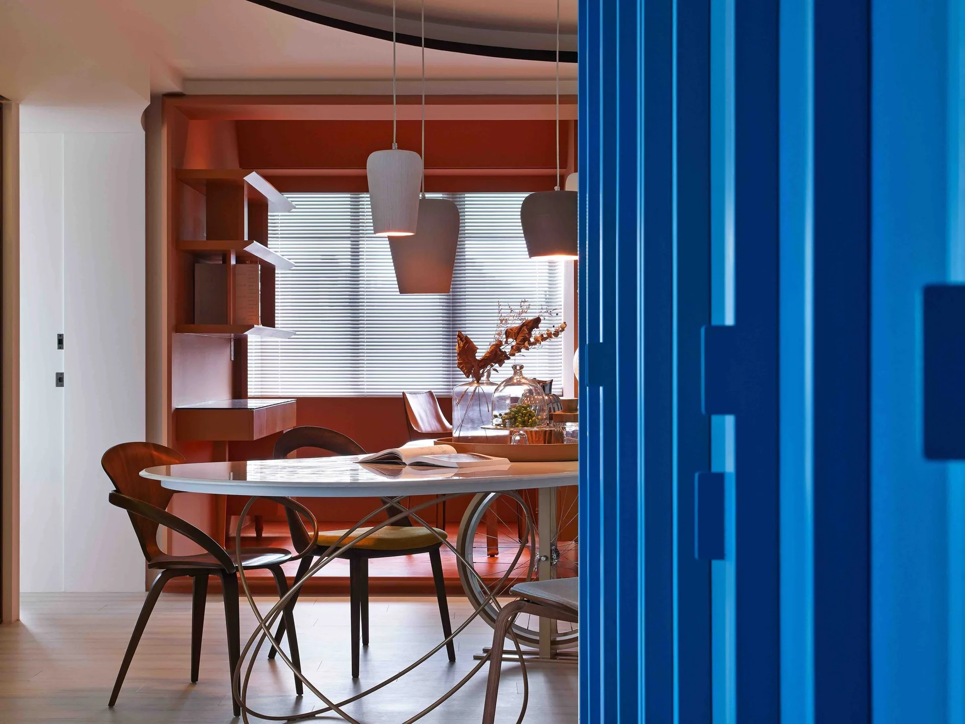 An oval dining table with wooden chairs is set against a terracotta shelving unit, with a bright blue sliding door partially visible in the foreground.