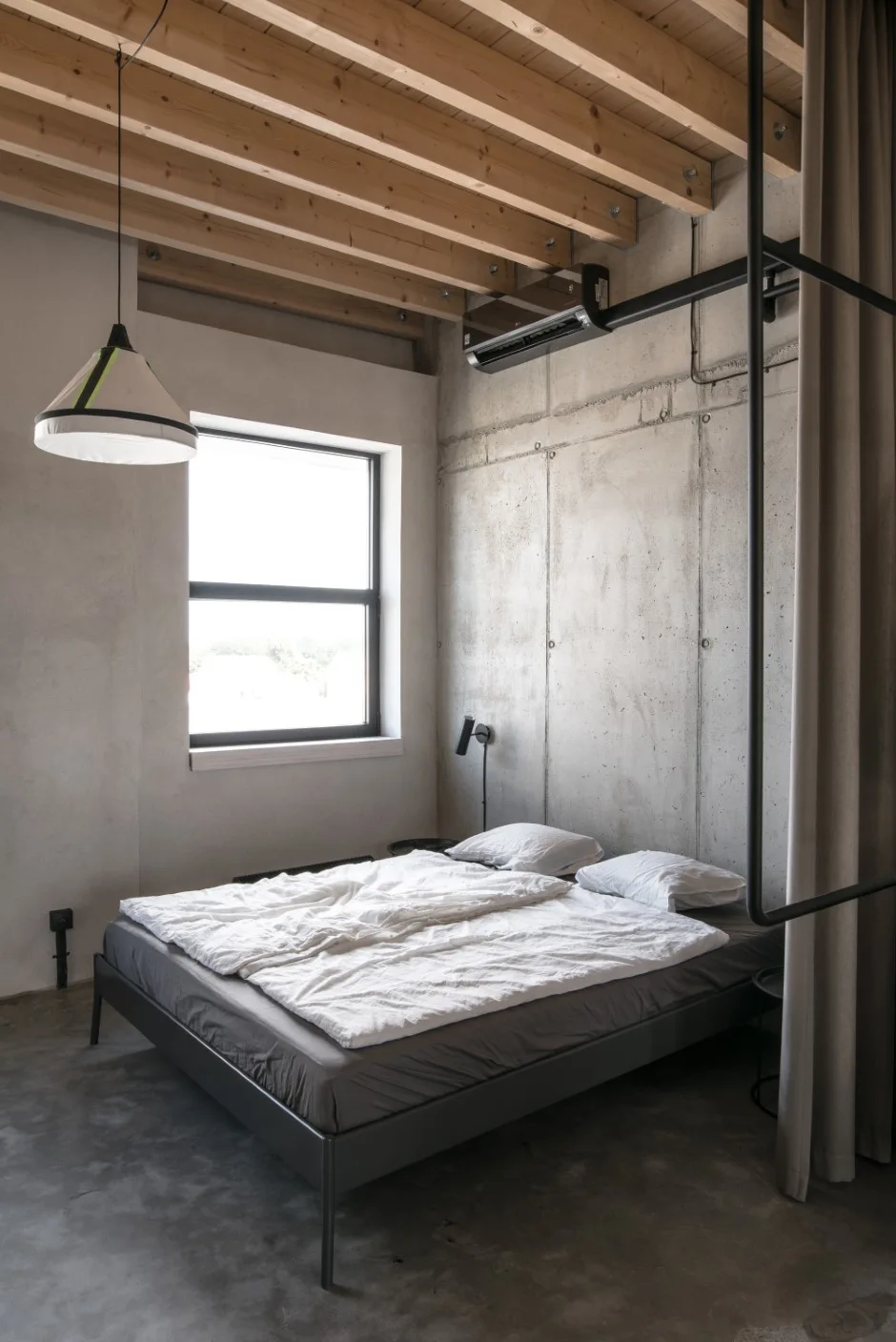 A minimalist bedroom features a gray metal bed against a raw concrete wall beneath an exposed wooden joist ceiling.