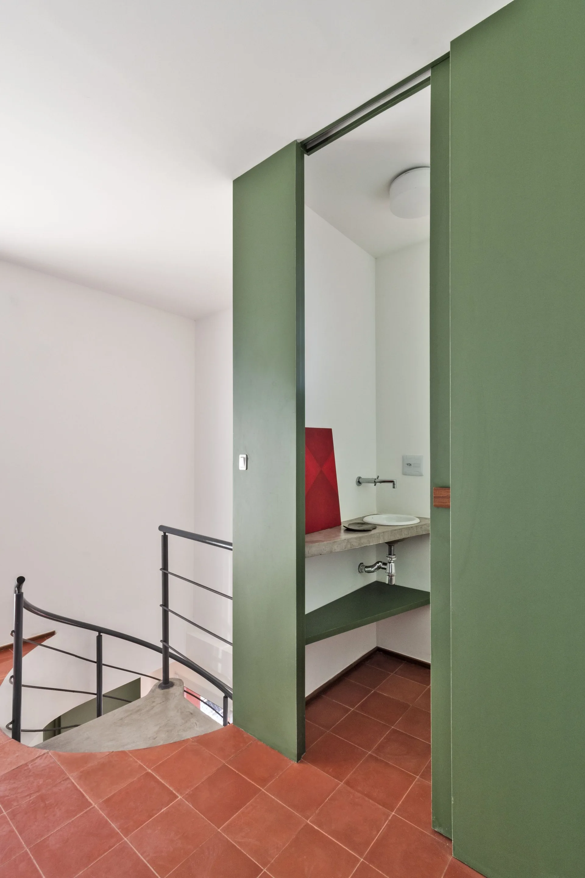 A modern powder room next to a spiral staircase features an olive green partition wall, a floating concrete sink counter, and terracotta tiled floors.