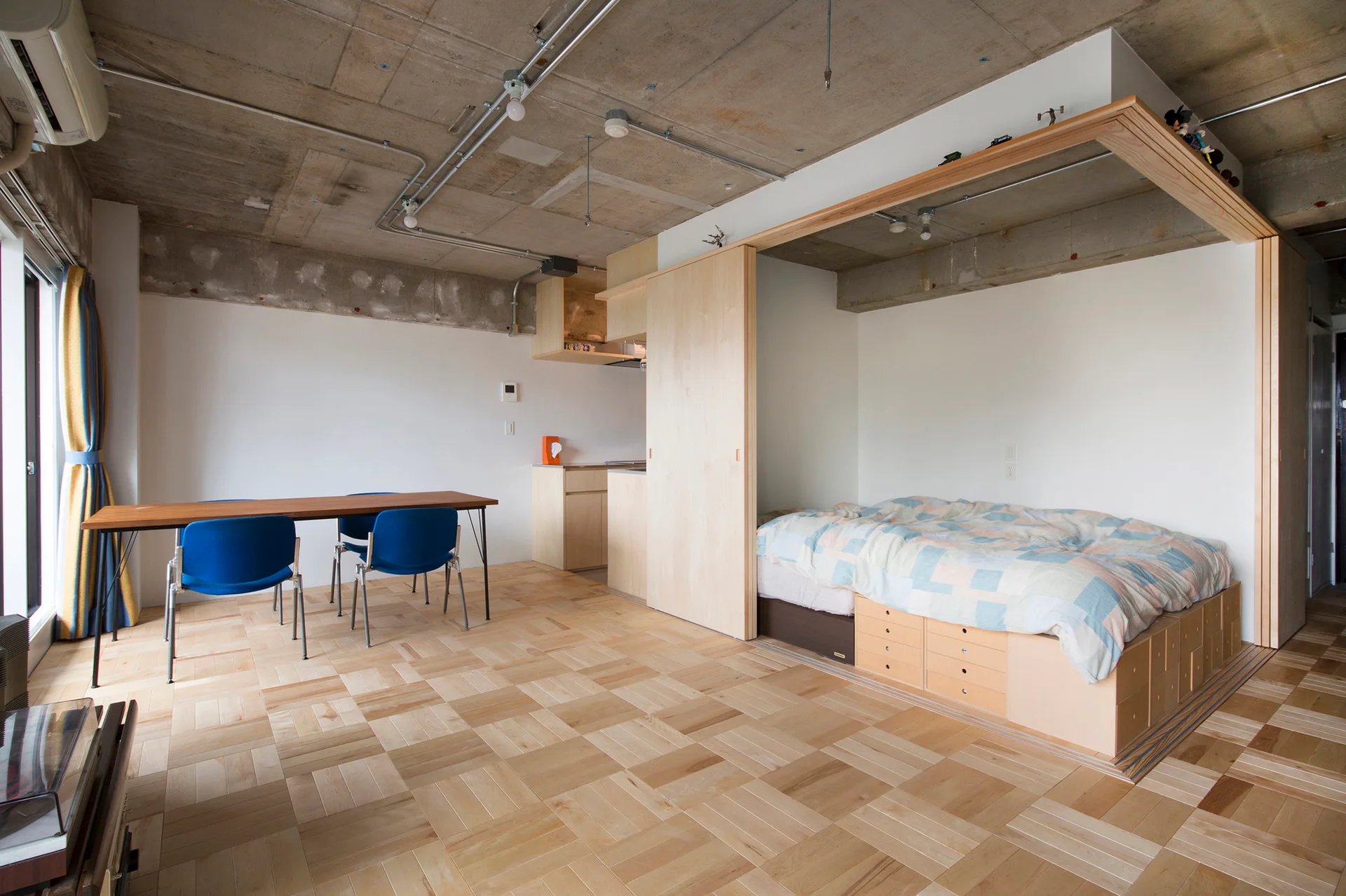 A studio apartment features a platform bed with built-in drawers, a light wood sliding partition, a dining table, and an exposed concrete ceiling.