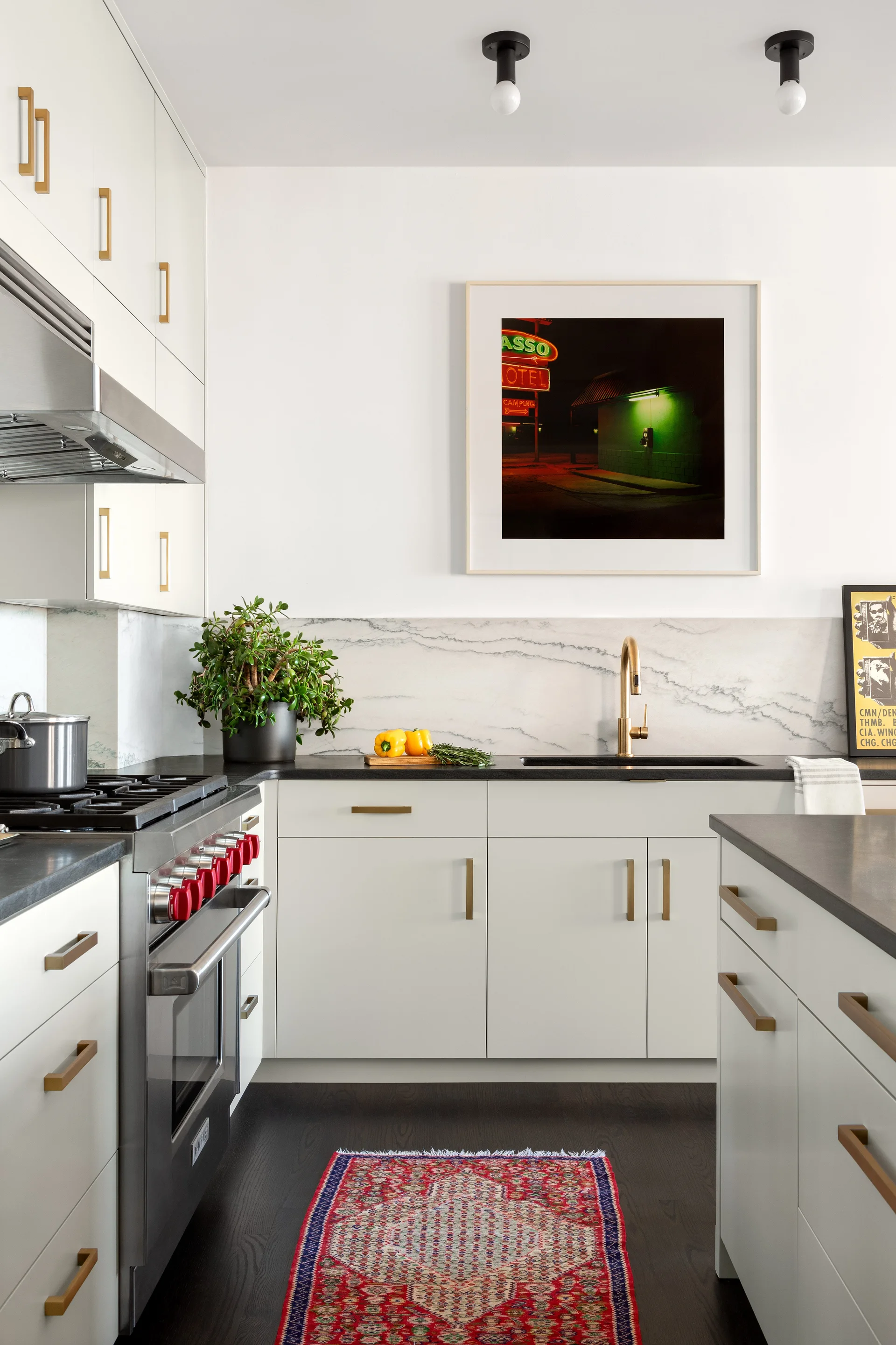 A modern kitchen featuring white cabinets with gold pulls, a marble backsplash, dark countertops, and a patterned red runner rug on dark wood floors.