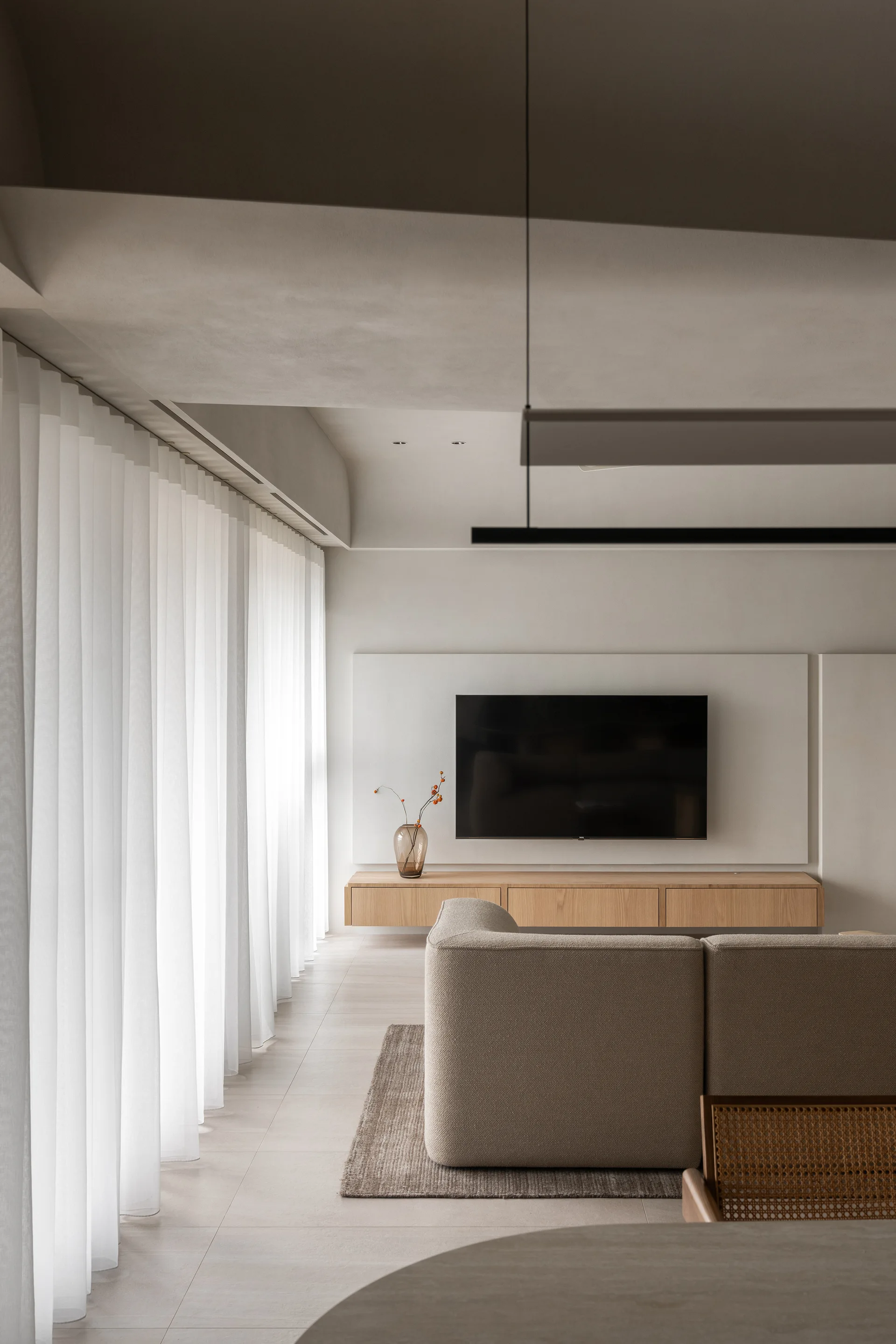 A minimalist living room featuring a modular beige sofa, floor-to-ceiling sheer curtains, and a floating wood console beneath a wall-mounted TV.