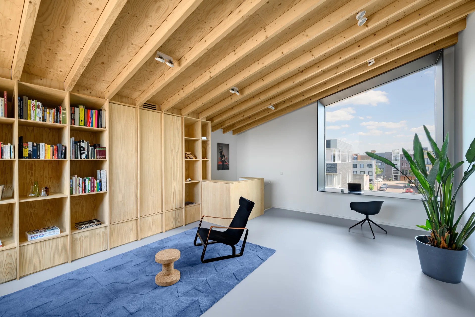 A minimalist room featuring a sloped plywood ceiling, a full-wall wooden bookshelf, a blue area rug, and a large window overlooking the street.