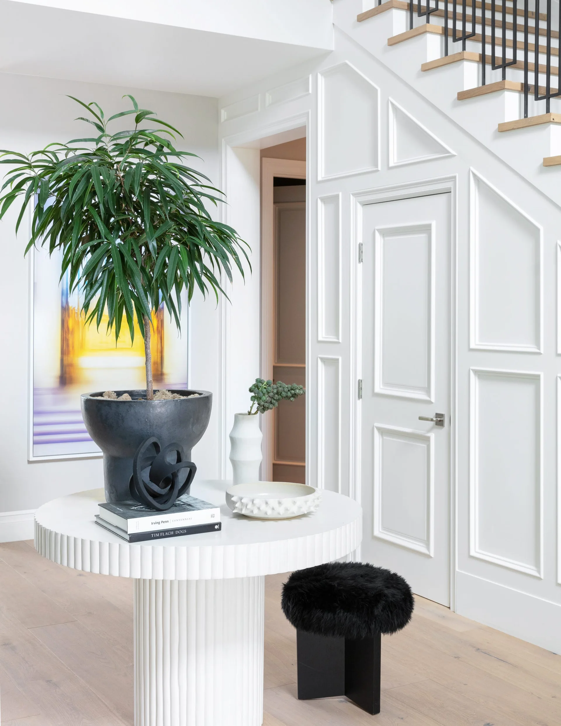 A bright entryway features a fluted white pedestal table with a large potted plant next to a staircase and white paneled walls.