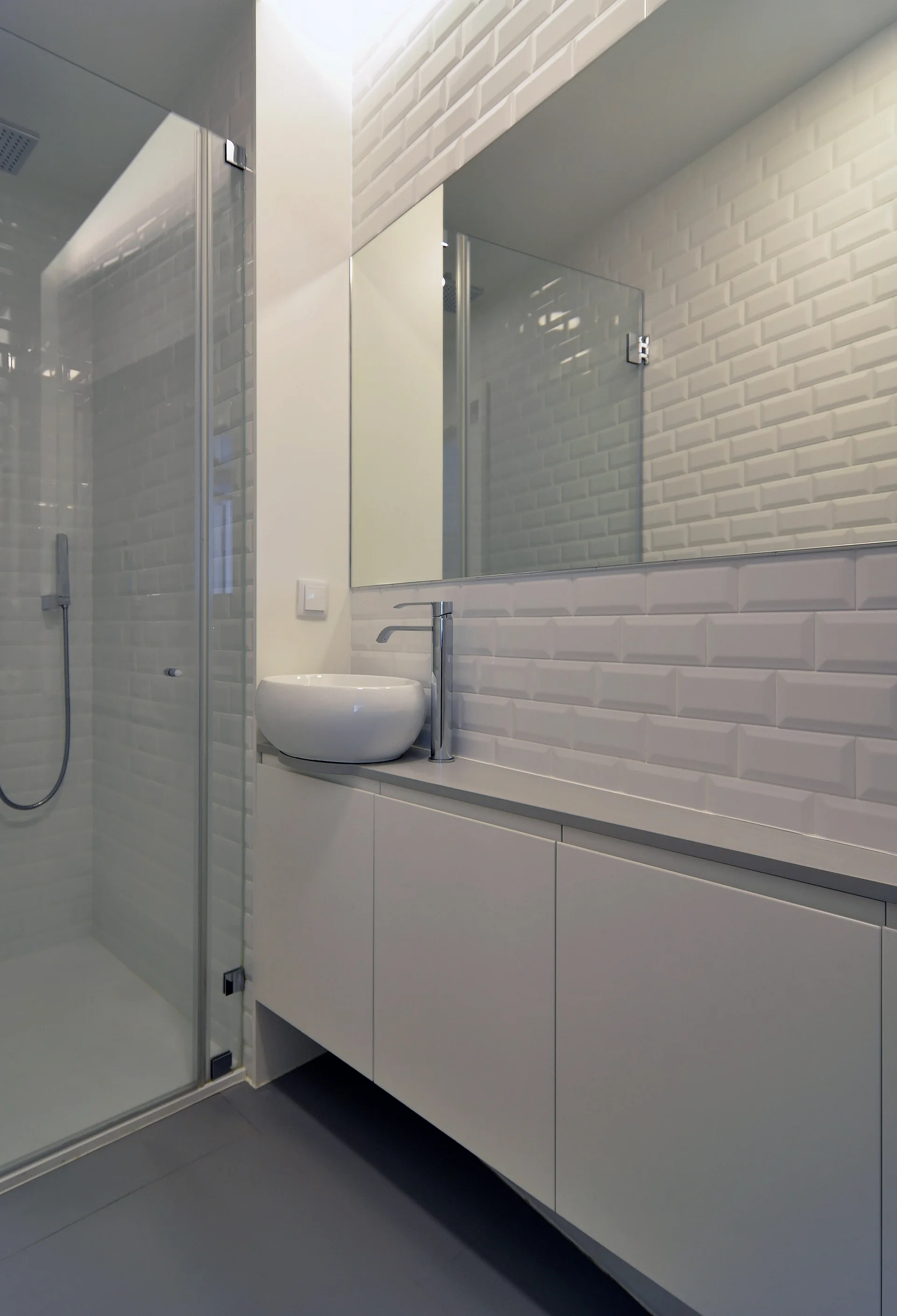 A minimalist bathroom features a white floating vanity with a vessel sink, a large mirror, and glossy subway tiles covering the walls.