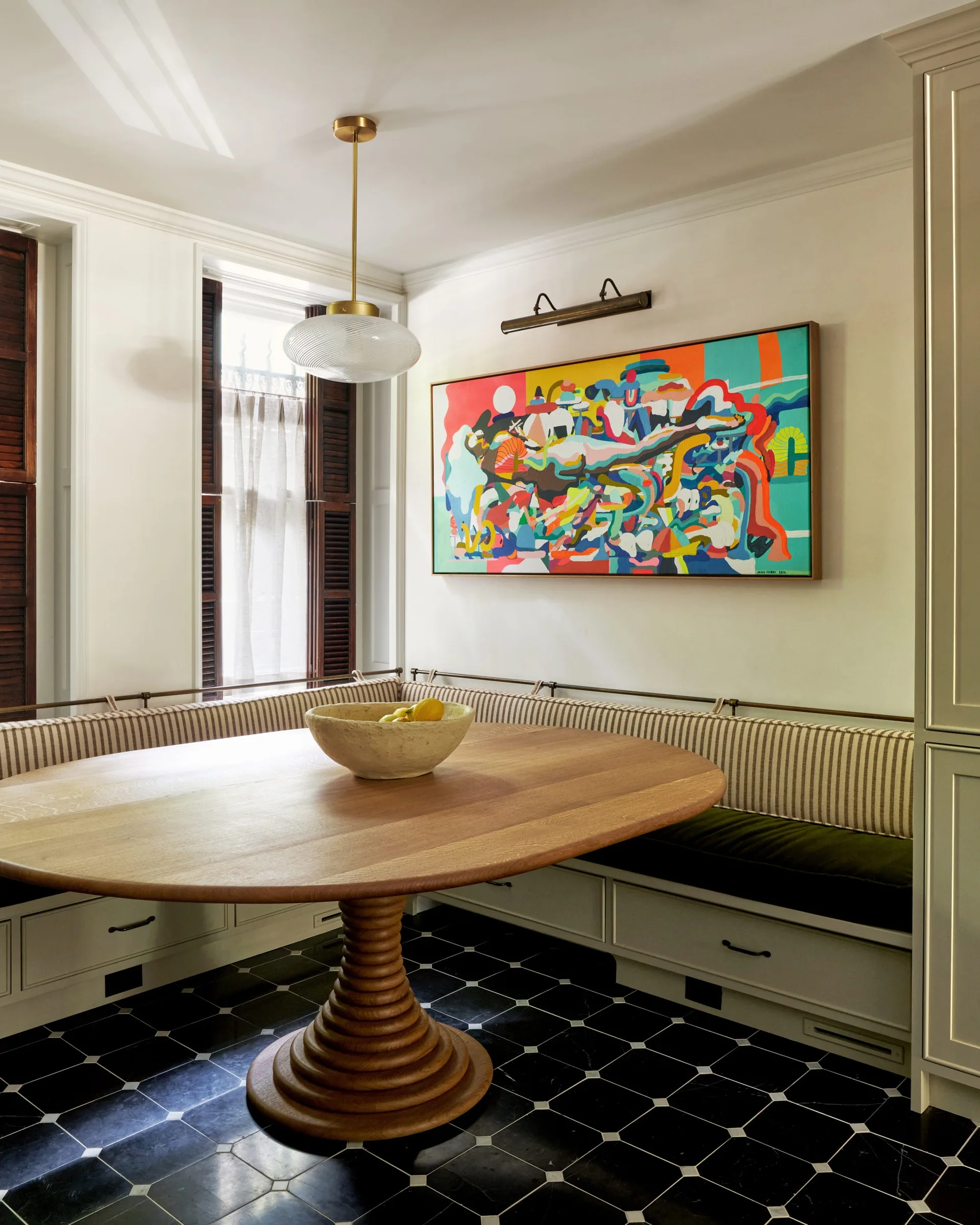 A dining nook features a round wooden pedestal table, built-in banquette seating with striped cushions, checkered tile floors, and a vibrant painting.