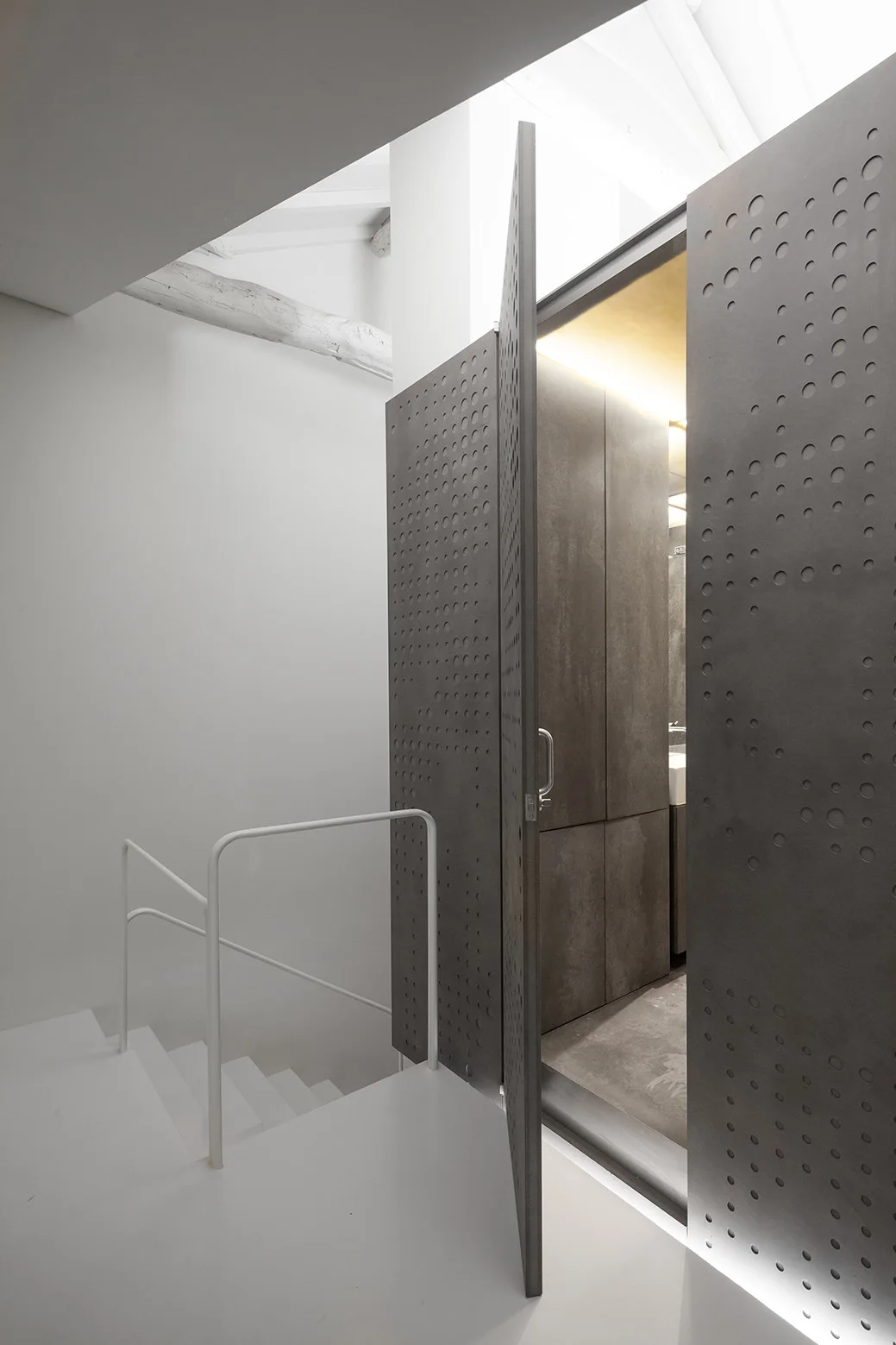 A minimalist white staircase with a white handrail stands next to a large, charcoal-gray perforated metal pivot door glowing with warm light.