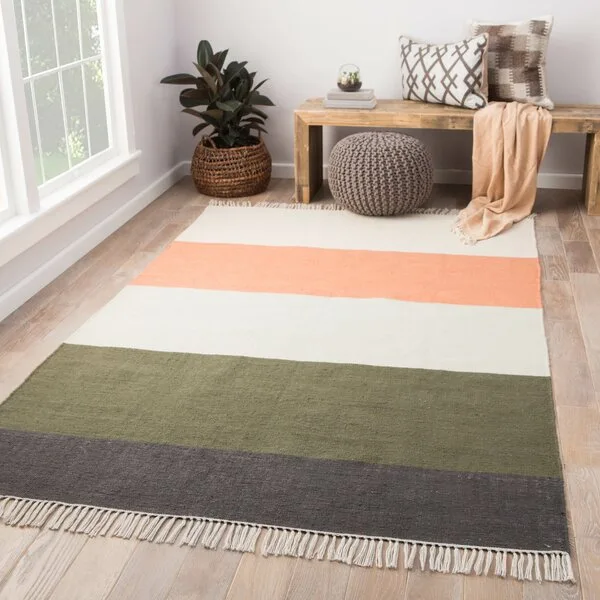 A multi-colored striped flatweave area rug sits on a light wood floor next to a rustic wooden bench, a gray knitted pouf, and a potted plant.