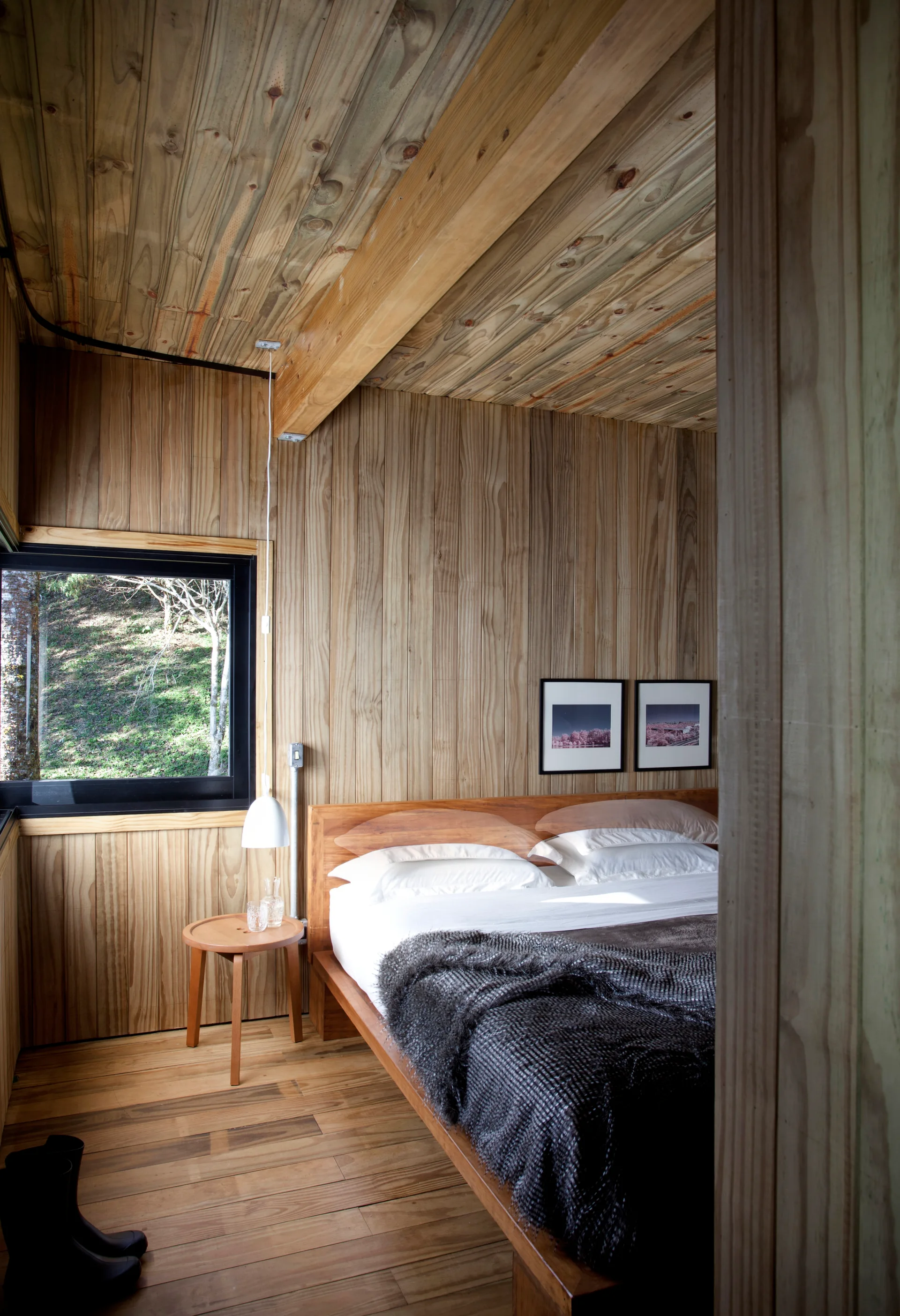 A minimalist bedroom featuring vertical wood-paneled walls, a matching wood ceiling, a wooden bed with white bedding, and a black-framed window.