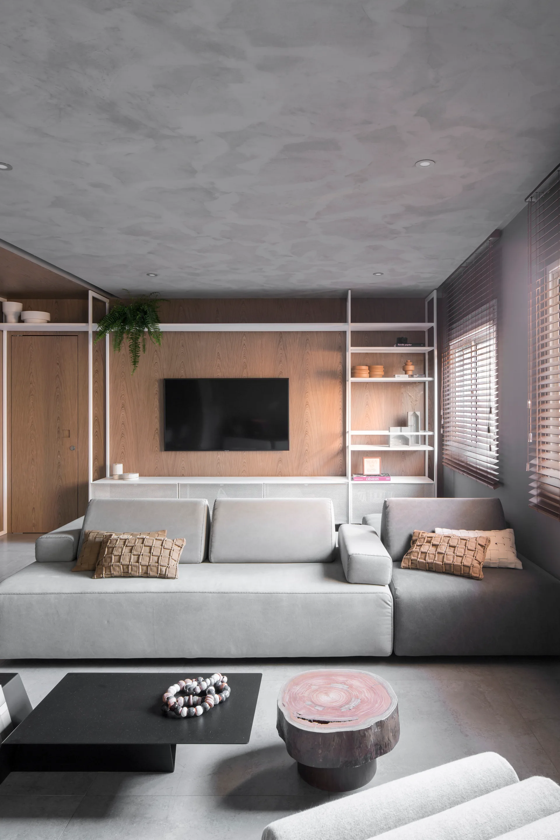 A grey sectional sofa sits in front of a wood-paneled wall with a mounted TV, white metal shelving, and a grey textured ceiling.
