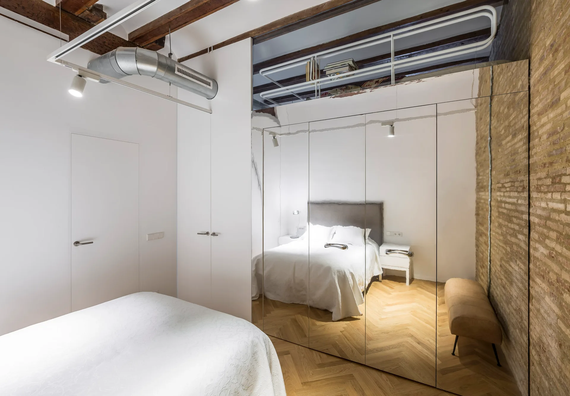 A loft bedroom featuring a floor-to-ceiling mirrored wardrobe reflecting a bed, set against an exposed brick wall and herringbone wood floors.
