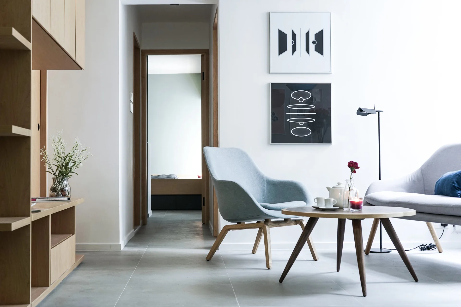 A minimalist living room features a light blue armchair, a round wood coffee table, and stacked geometric artwork on a white wall.