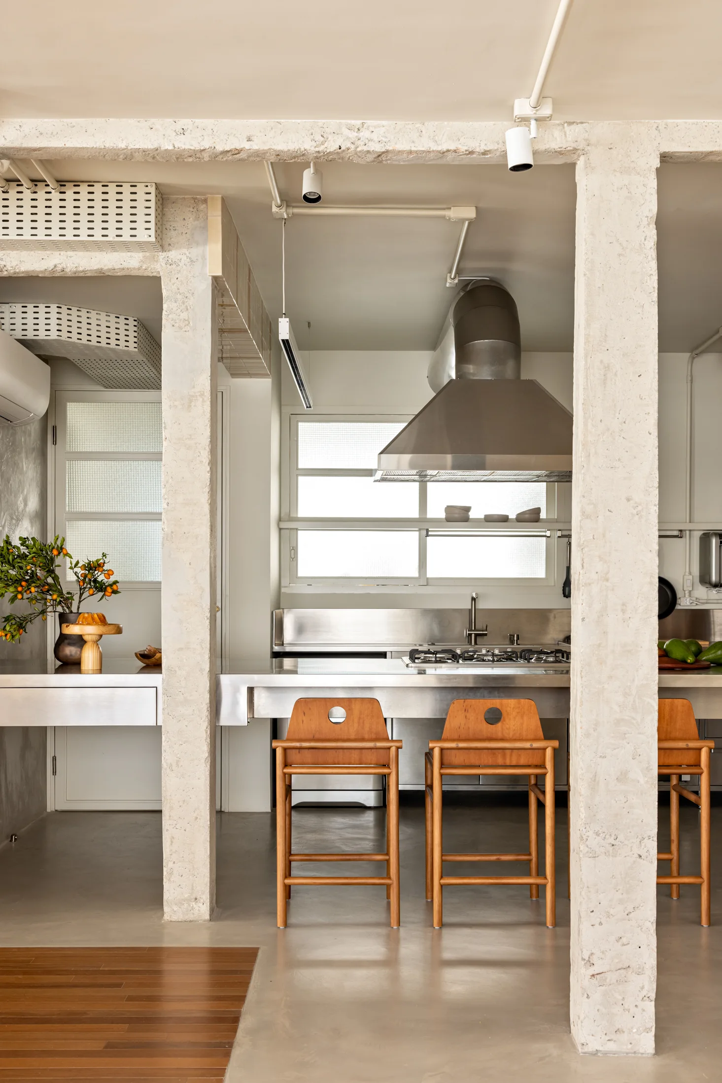 Three wooden bar stools sit at a stainless steel kitchen island framed by rough, exposed concrete support columns.