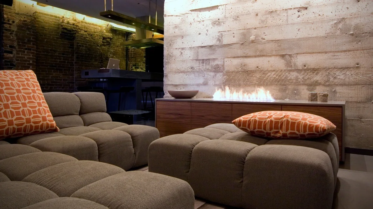 A modern living room features a tufted modular sofa with orange pillows facing a linear fireplace set against a textured board-formed concrete wall.
