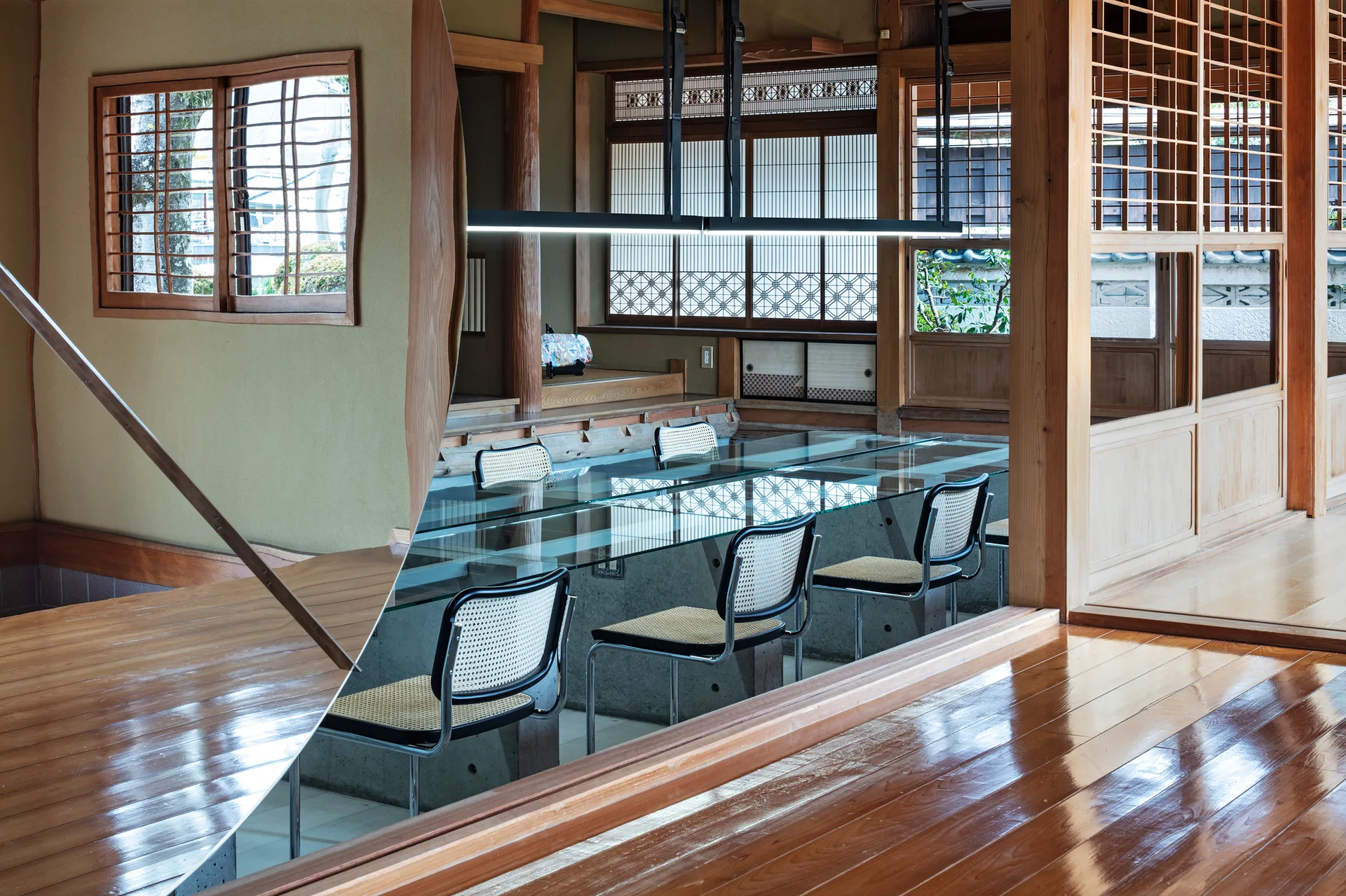 A modern dining room featuring a long glass table with cane-back chairs surrounded by traditional Japanese wood columns and sliding screen doors.