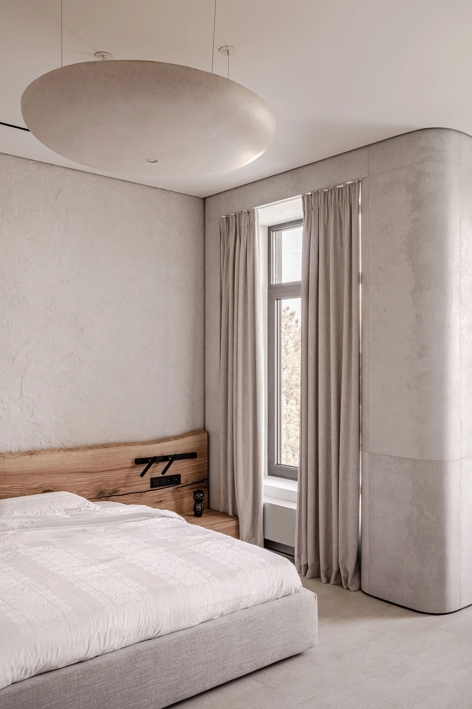 A minimalist bedroom featuring a low upholstered bed with a custom wood headboard against textured concrete plaster walls.